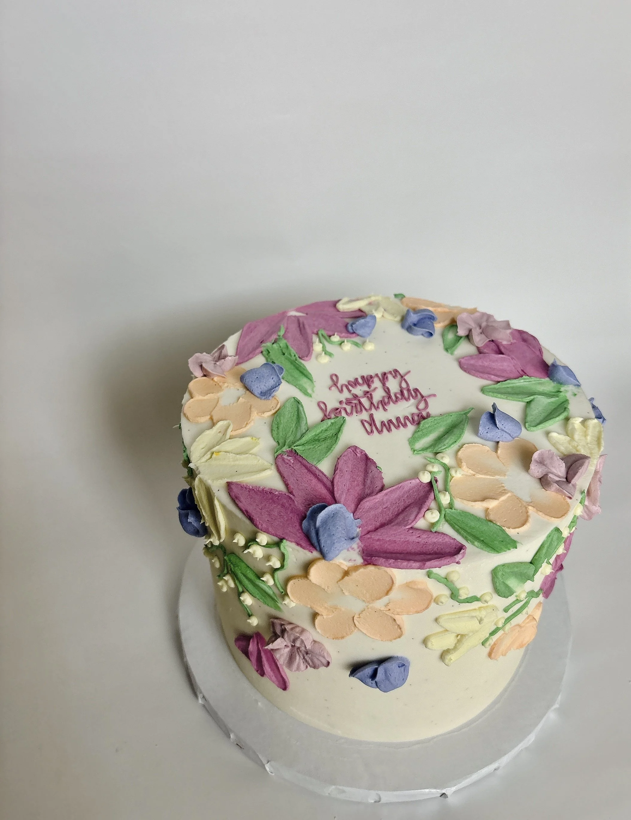 A white round cake decorated with colorful buttercream flowers and leaves, with the words "Happy Birthday" written in pink on top.