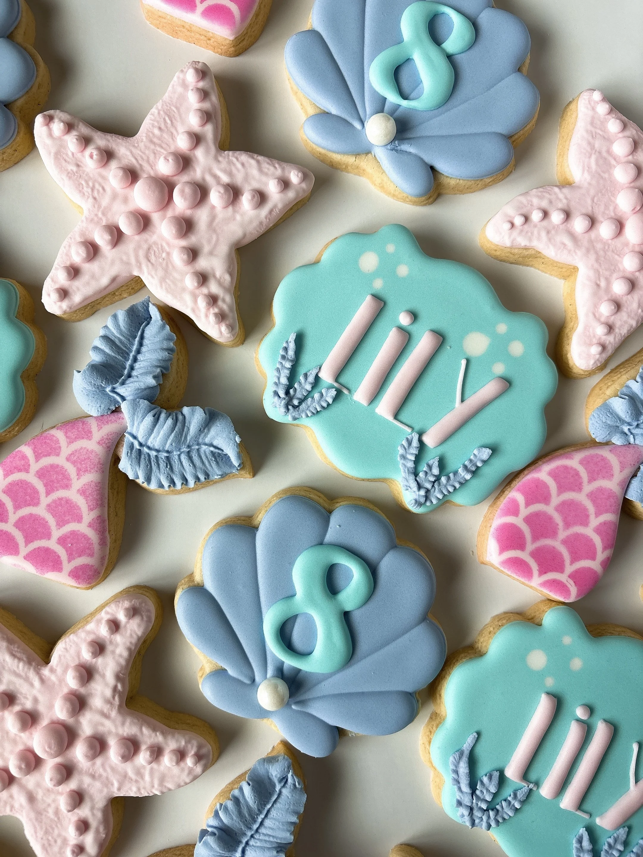 Colorful decorated cookies with pastel icing in shapes like stars, clouds, hearts, and leaves, some with the word 'Hi' on them, arranged on a white surface.