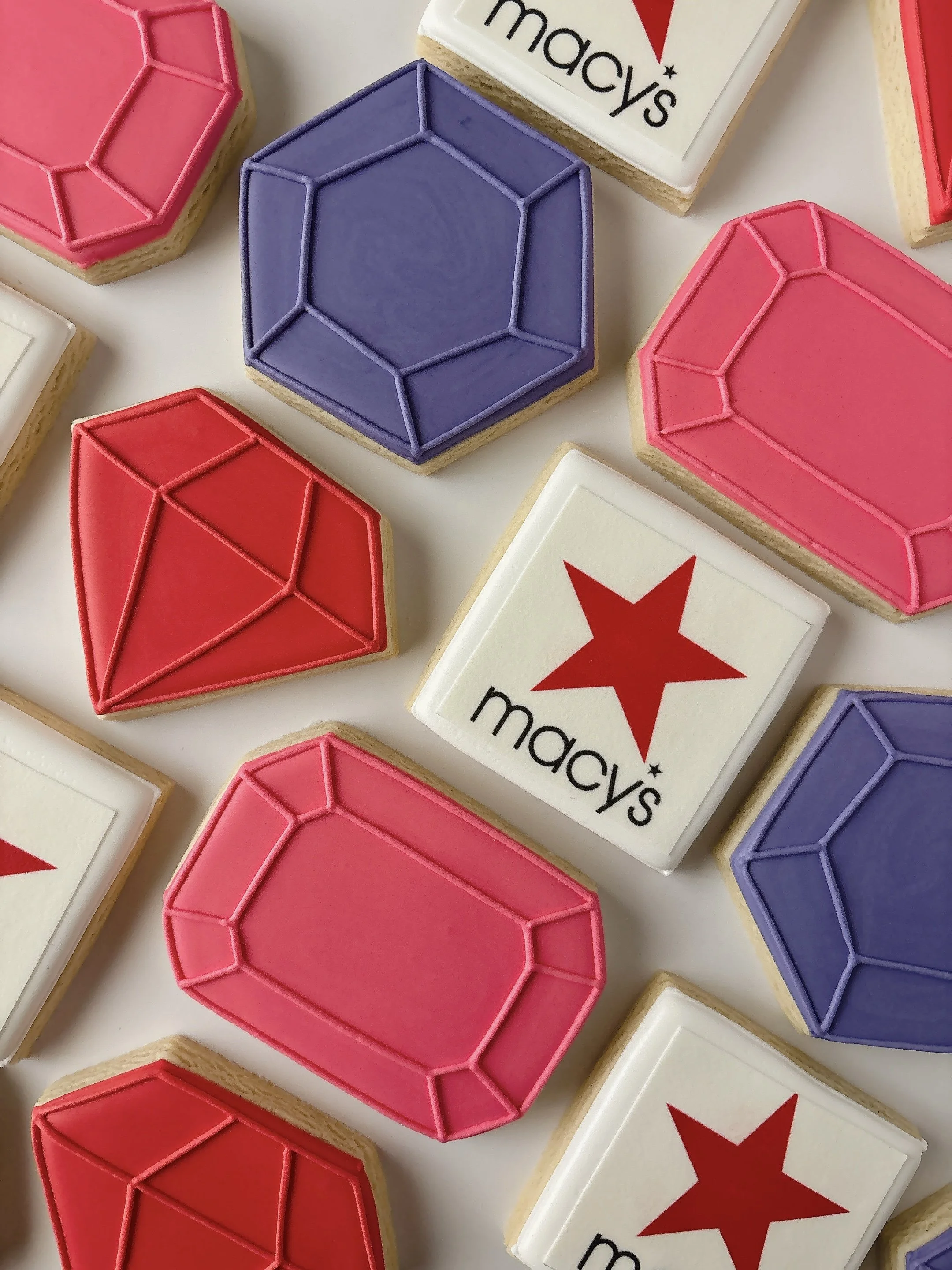 Decorative sugar cookies shaped like hearts and rectangles with pink, purple, and white icing, some with a red star and 'macy's' written on the white ones.
