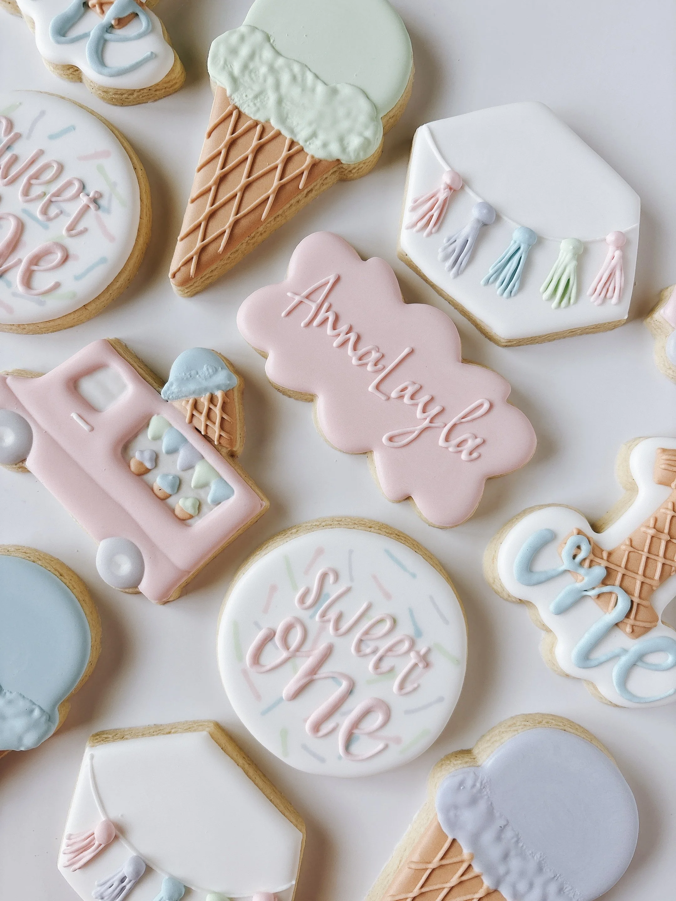 Decorated cookies with pastel colors and inscriptions, including an ice cream cone, cloud-shaped cookie with 'Ahnaya' written, and other birthday-themed cookies.