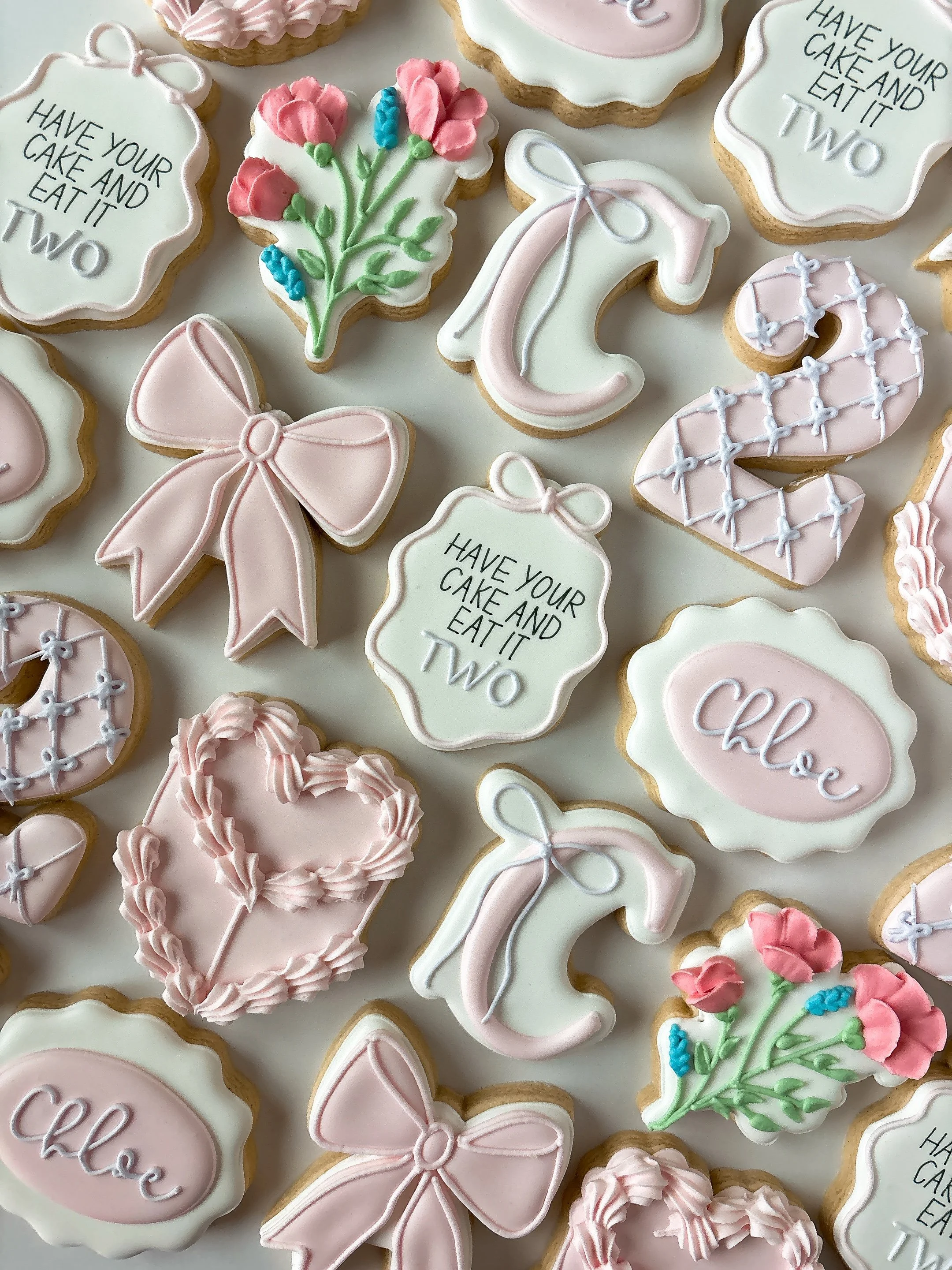 Assorted decorated cookies with pink, white, and green icing, shaped like bows, flowers, and speech bubbles. Some cookies feature messages such as "Have your cake and eat it two" and "Chloe."