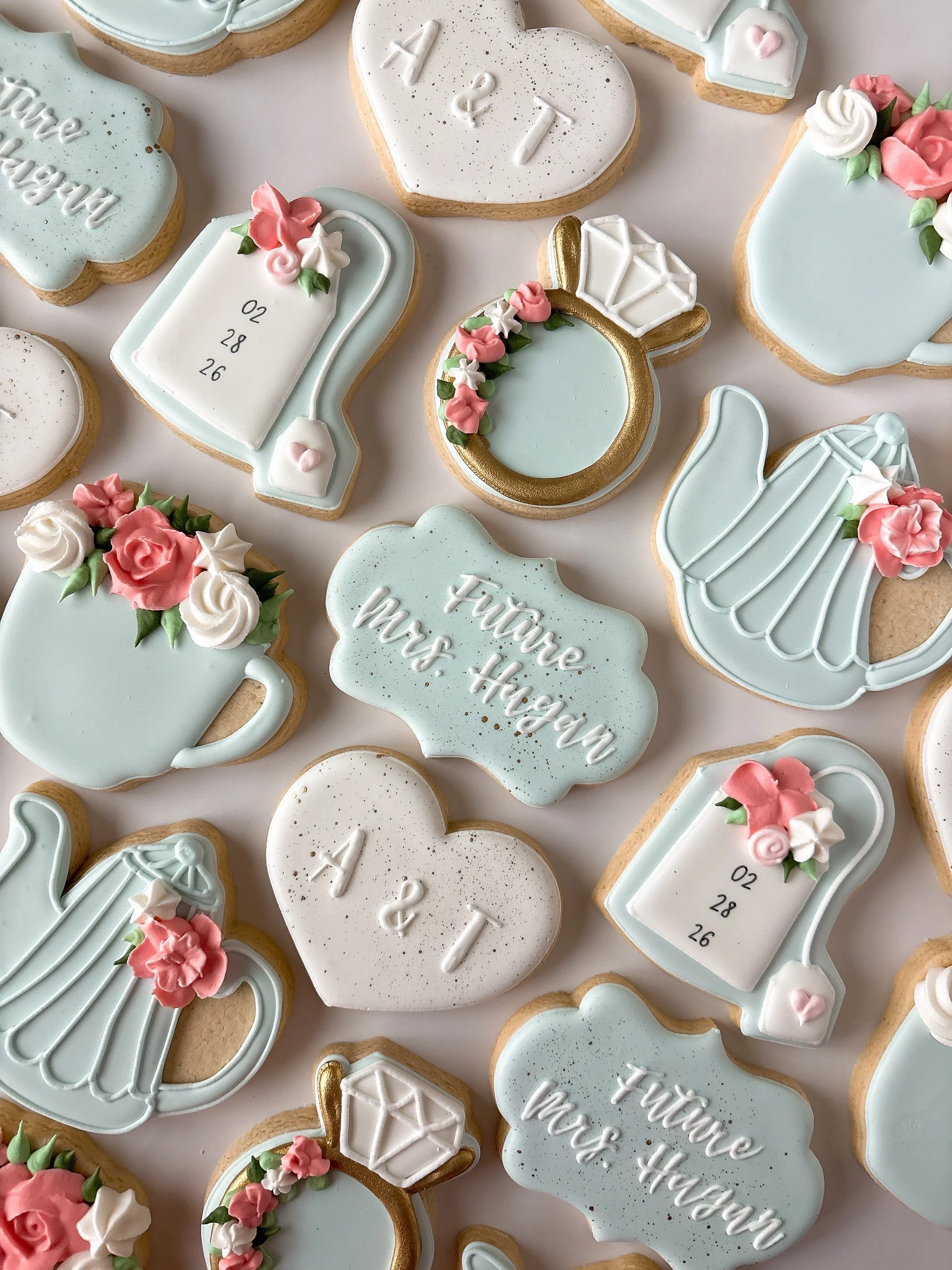 Decorative cookies with pastel blue, white, and pink icing, some with floral designs, a diamond ring, and text such as "Future Mrs. Hagan", "A & T" and a calendar date "02 28 26".
