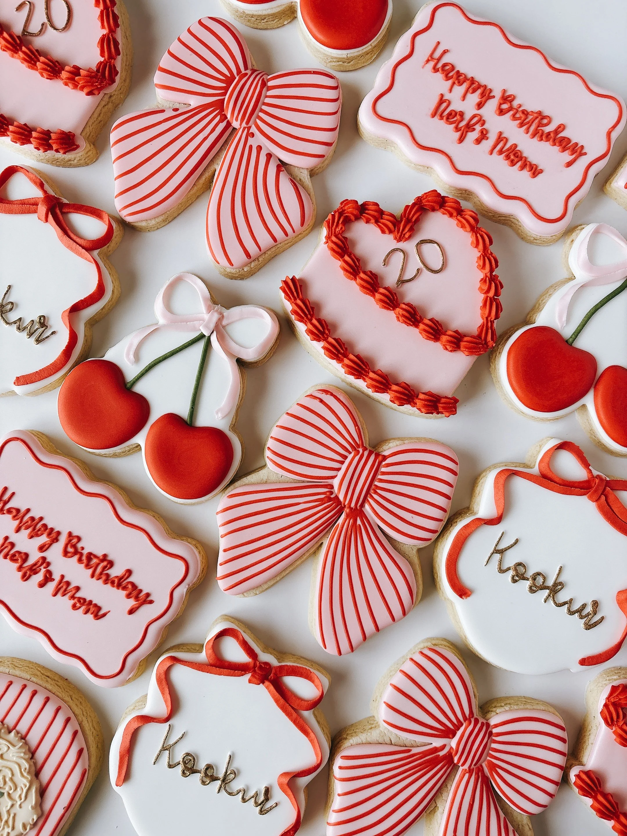 Collection of decorated cookies with red and white themes, some shaped like hearts, bows, and rectangles. Some cookies have birthday messages, including 'Happy Birthday Neri & Mony' and 'Kashoush' written in red, with various icing patterns and ribbo