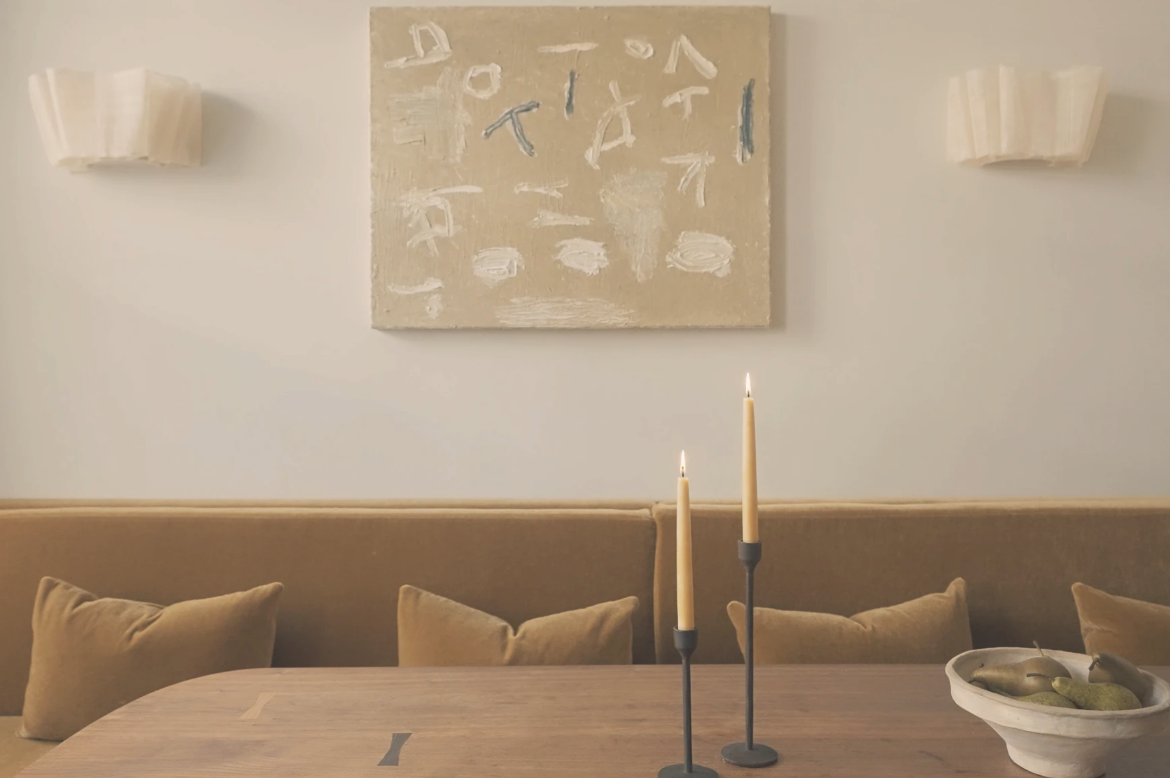 A living room with beige sofas, a wooden dining table, a wall-mounted beige canvas with white and gray abstract writing, two white wall sconces, and a beige and gray candle holder with three lit candles in the foreground.