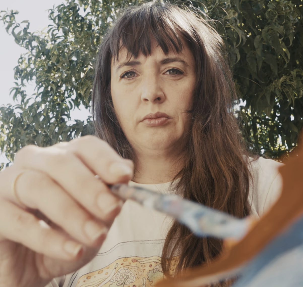 Artist, Billie Justice-Thompson, is outdoors, holding a paintbrush, painting.