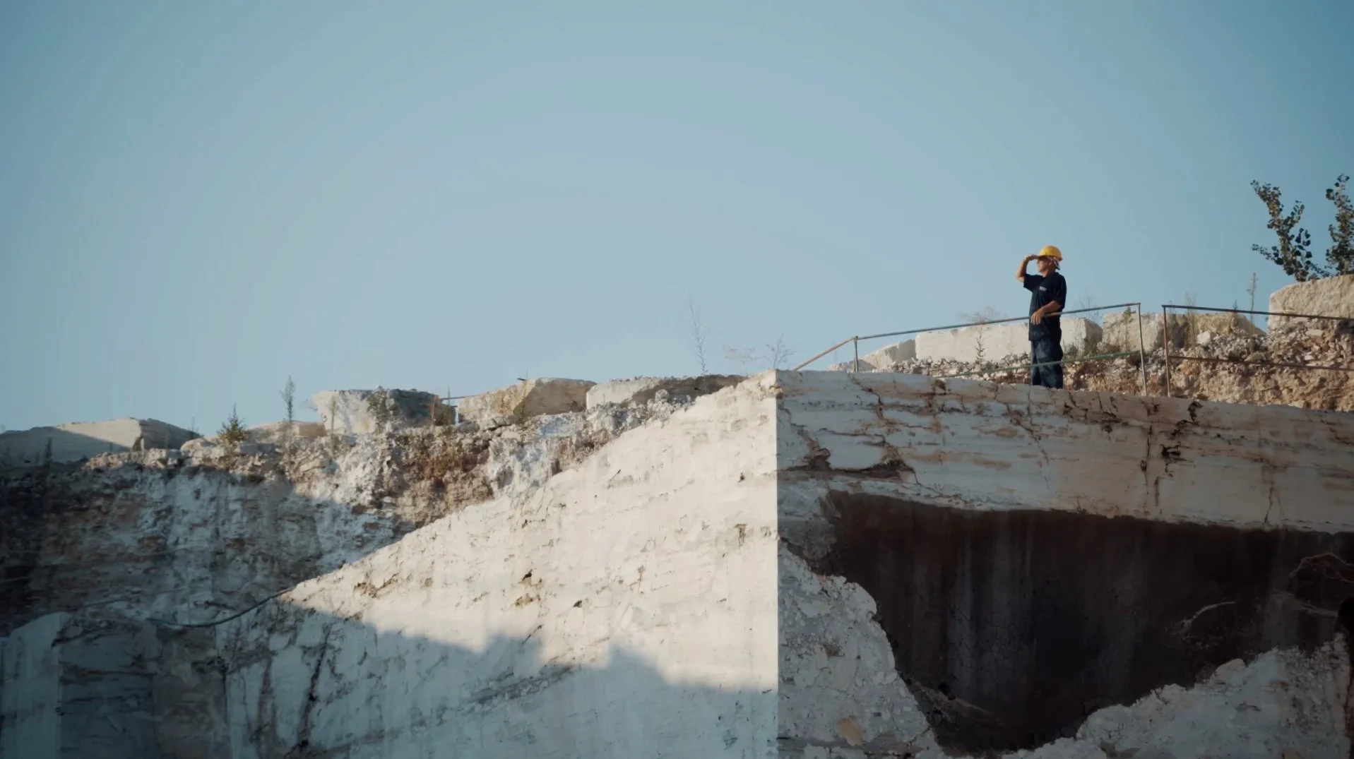 A person wearing a yellow hat and dark clothing standing on a rocky ledge, looking out over a landscape of white rock formations and sparse vegetation under a clear blue sky.