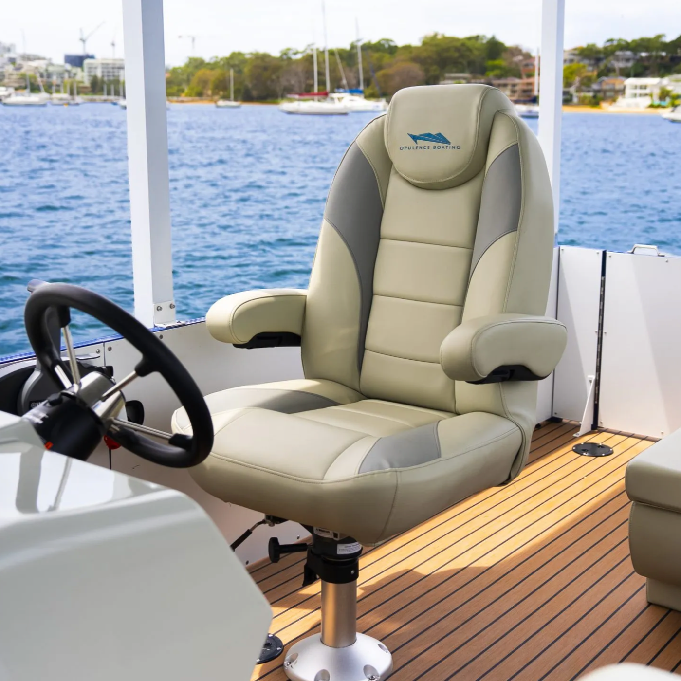 A captain's seat on a boat with a beige leather upholstery, featuring the logo 'Opulence Boating', situated on a wooden deck. The background shows calm water with sailboats and a shoreline with trees and buildings.