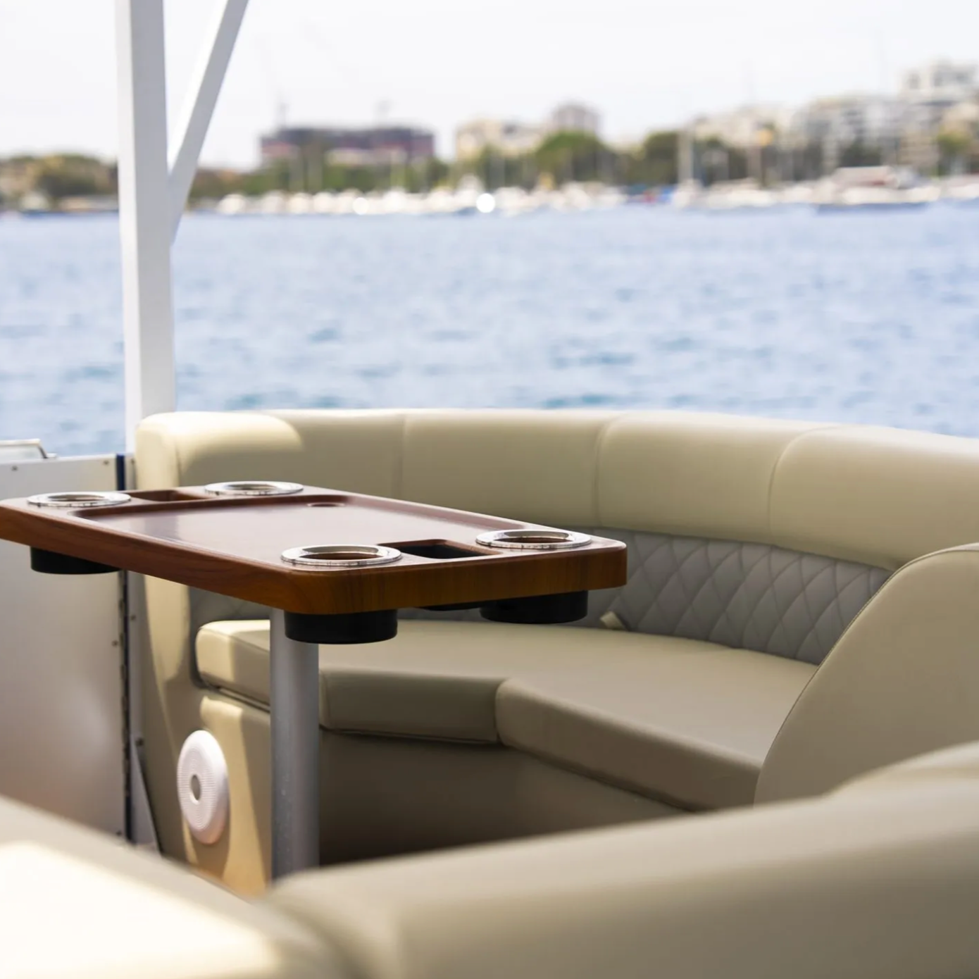 Inside view of a boat with a seating area, a wooden table with holders for drinks, and a water backdrop with boats and buildings in the distance.