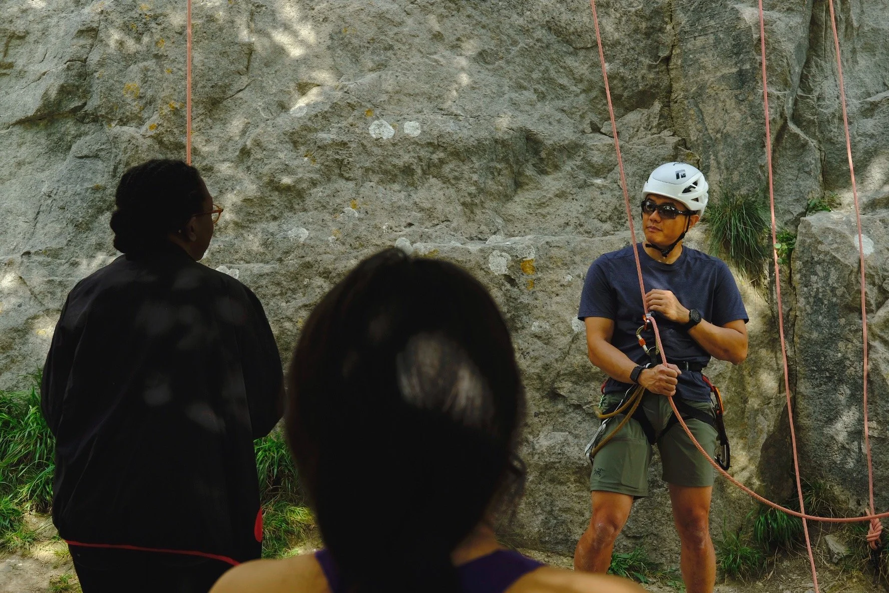 A person wearing a white climbing helmet, sunglasses, and a navy T-shirt giving instructions to a small group outdoors in front of a rock wall, with ropes attached for climbing.