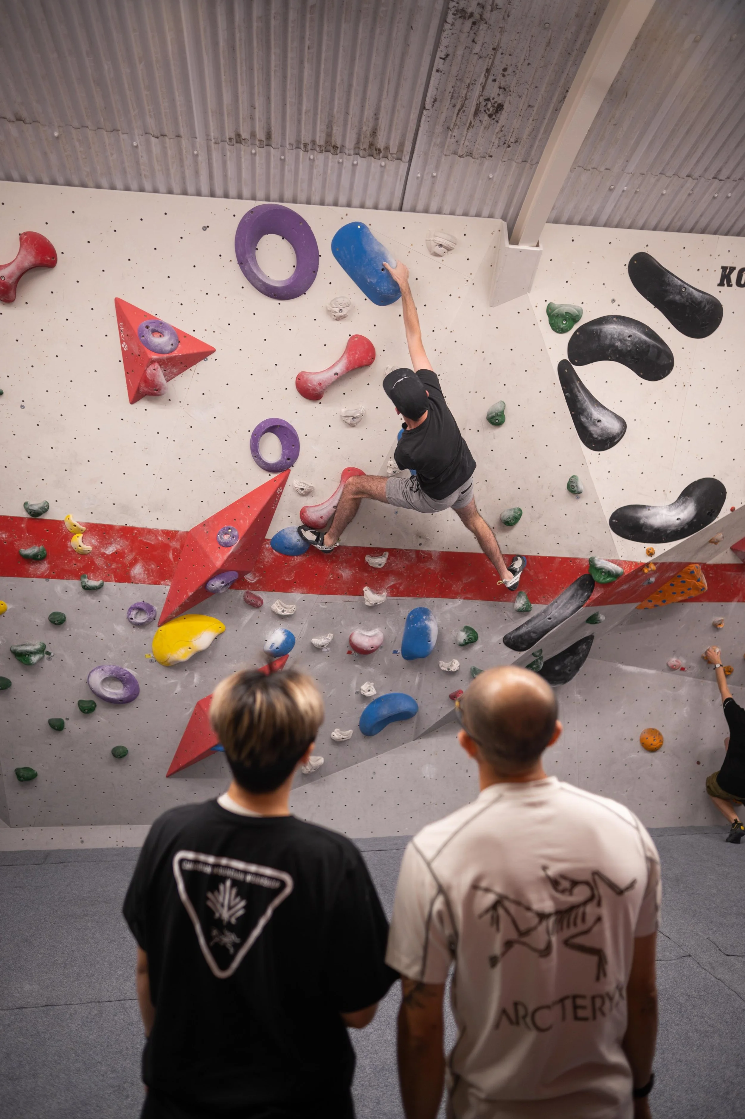 Indoor boulder climbing gym with colorful holds and a climber reaching for a large blue hold, while two spectators watch from the foreground.