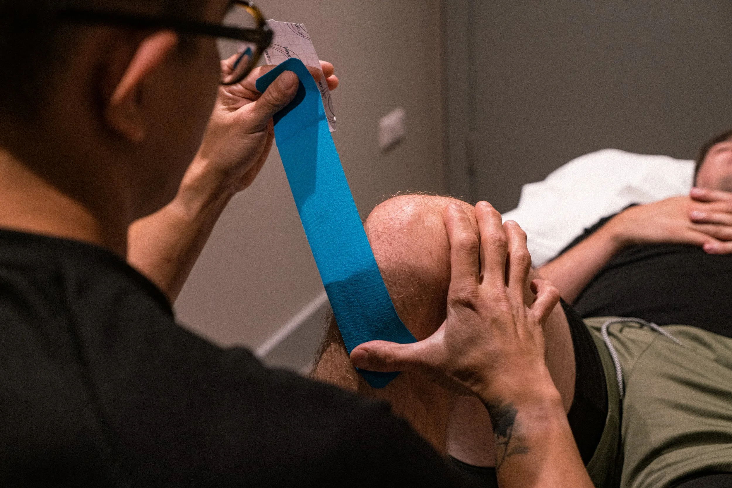 A person is holding a piece of paper with blue medical tape attached to their knee, while another person is supporting their head and shoulder during what appears to be a medical or emergency situation.