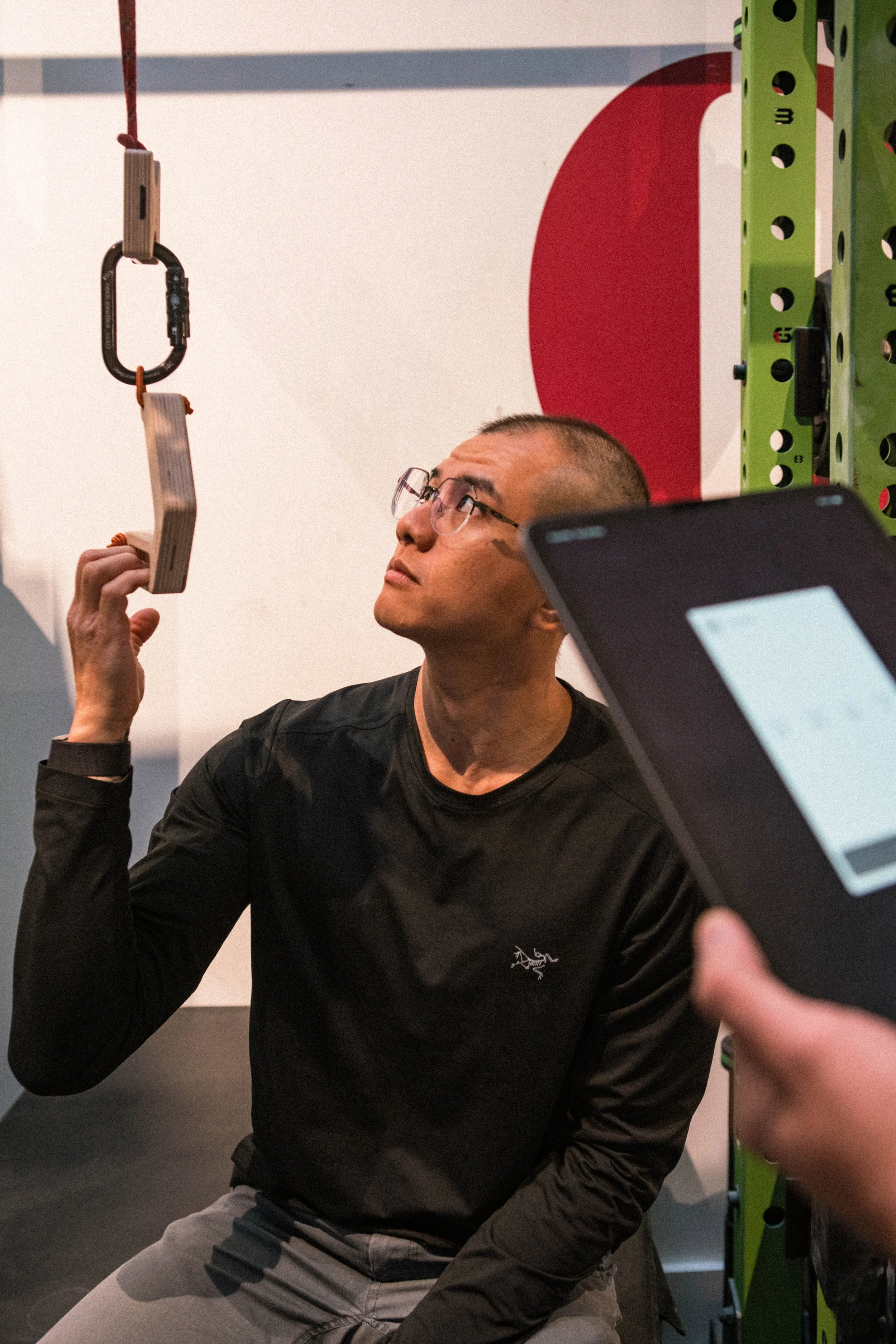 A man wearing glasses and a dark shirt is sitting on a chair, looking up at a hanging wooden workout tool with a black metal clip and a red strap. In the background, a green workout frame and a red and white wall are visible.
