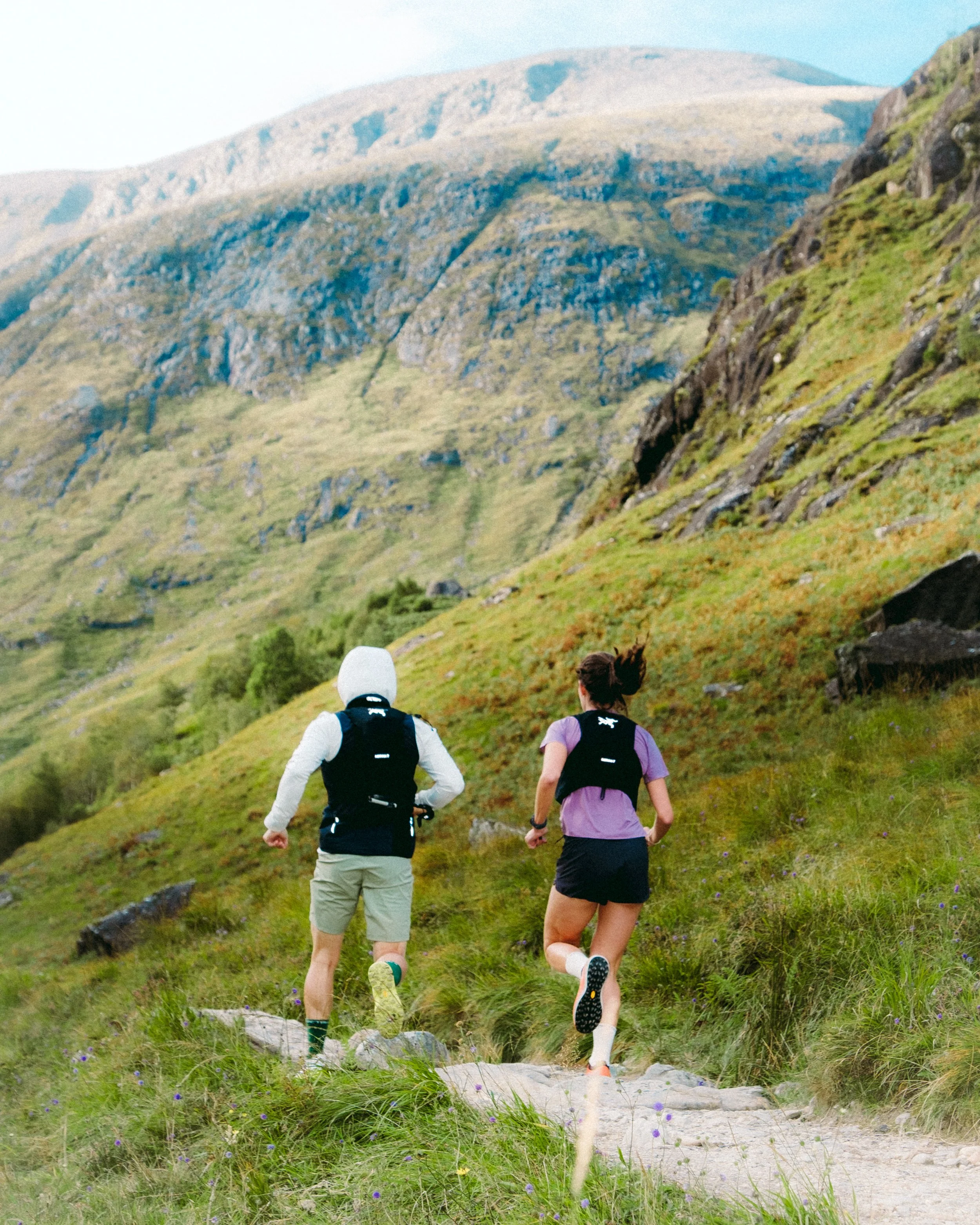 Two women trail running up a grassy mountain trail surrounded by green grass and wildflowers, with towering mountain peaks in the background.