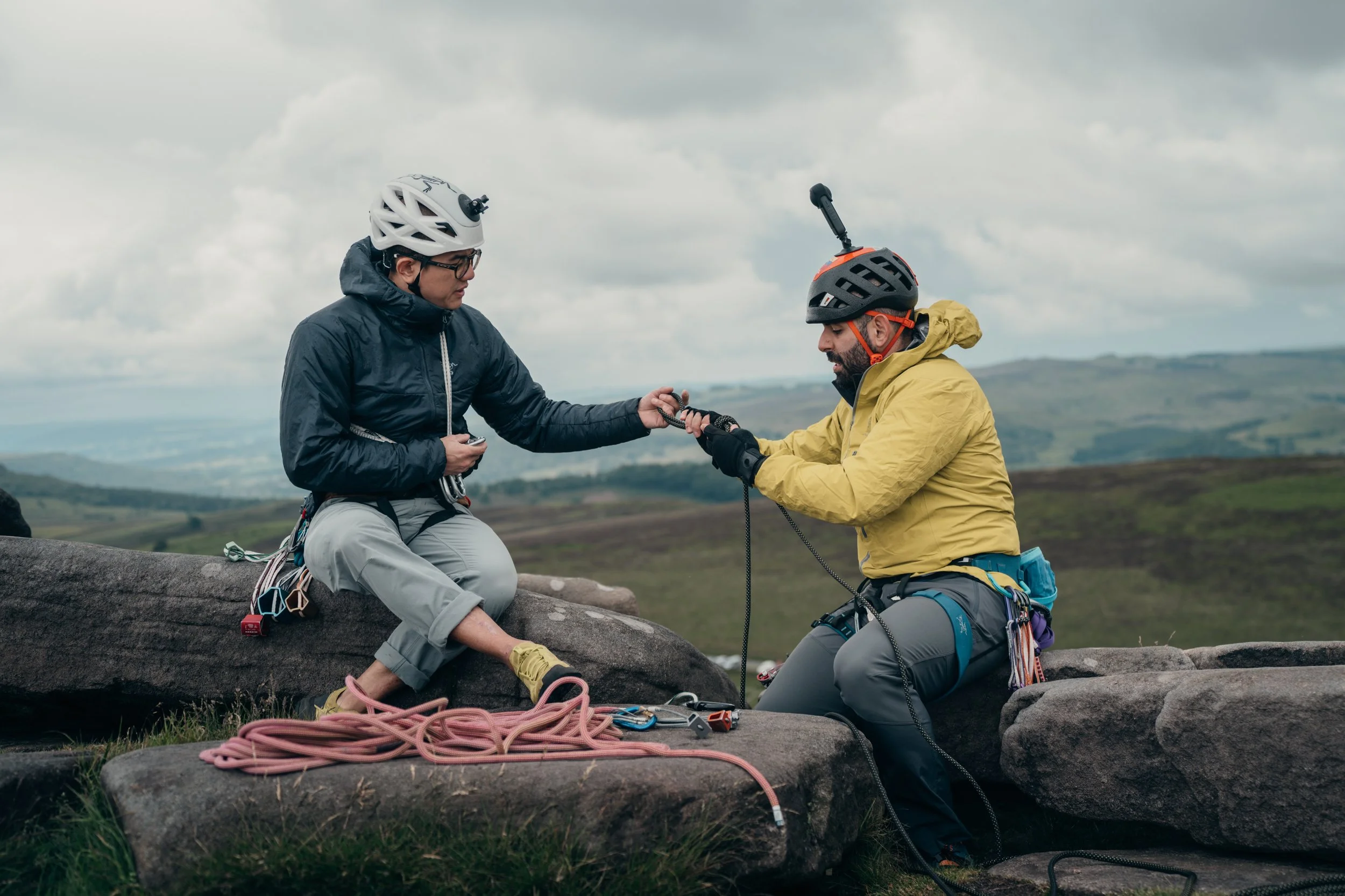 Two rock climbers, with helmets and gear, securing a climber sitting on a large rock outdoors during daytime with cloudy sky and rolling hills in the background.
