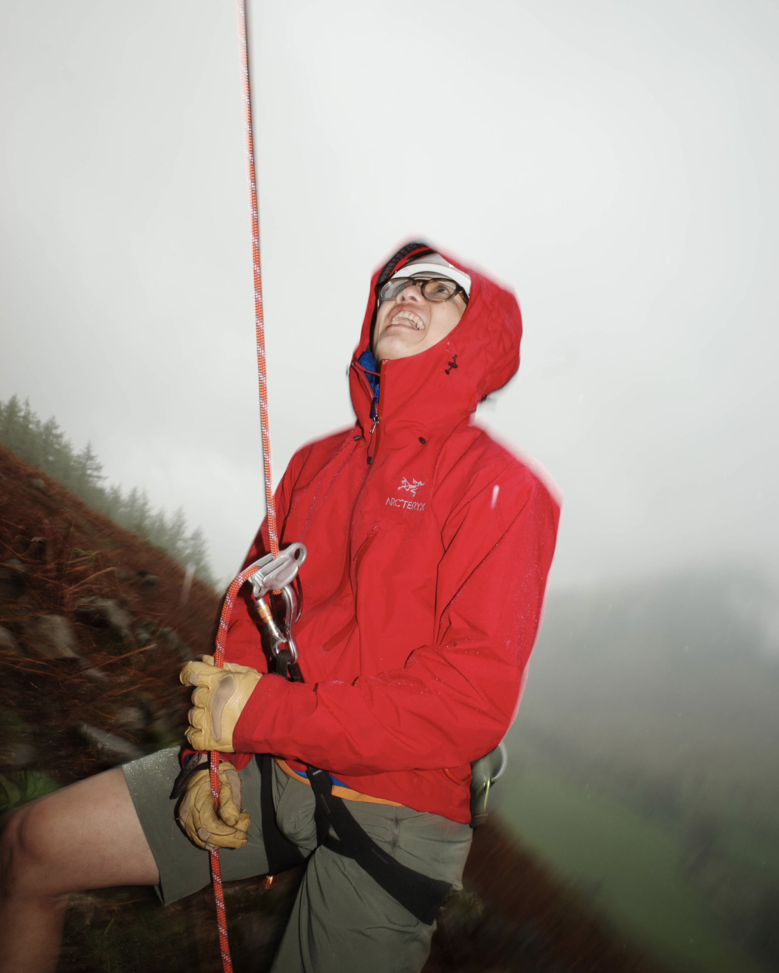 Person wearing a red Arc'teryx jacket and gloves, holding climbing gear, outdoors in a foggy, forested area.