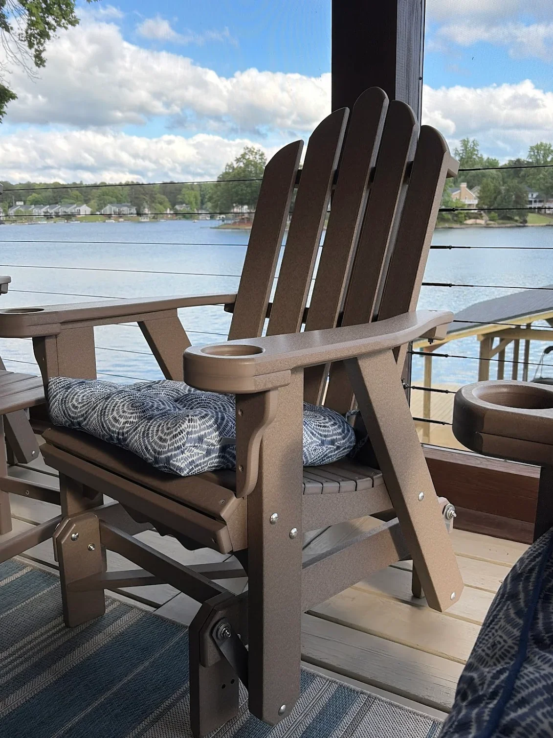 A porch overlooking a lake with Adirondack chairs, a cushion on the chair, and a view of houses across the water under a partly cloudy sky.