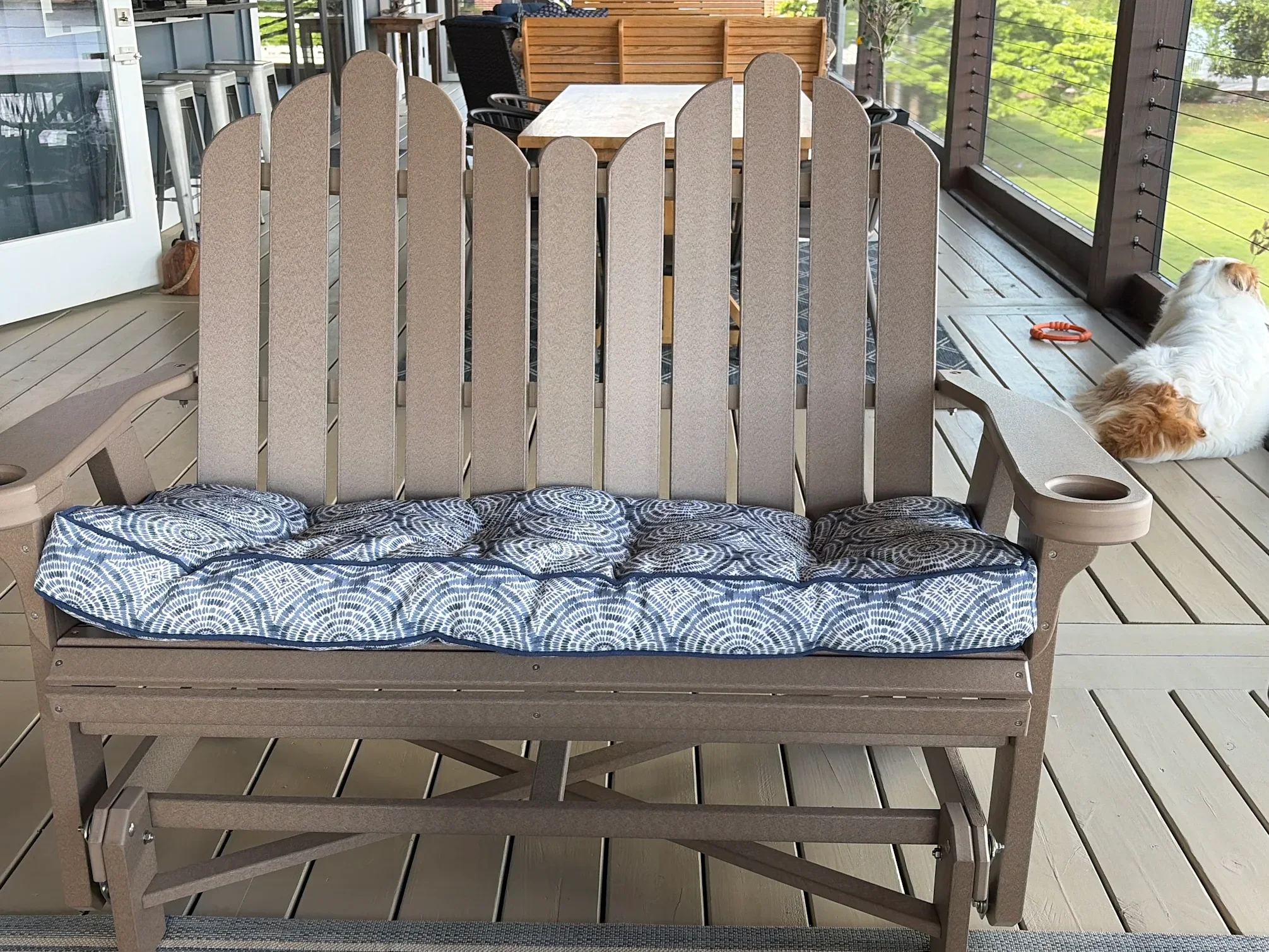 A screened porch with a wooden deck. In the foreground, a beige wooden glider with a blue patterned cushion on the seat. Behind it, a beige outdoor table with a white surface. To the left, a brown leather bag on the floor. To the right, a dog lying down on the deck with a bright orange toy beside it. The background shows lush green trees and a house beyond the screen.