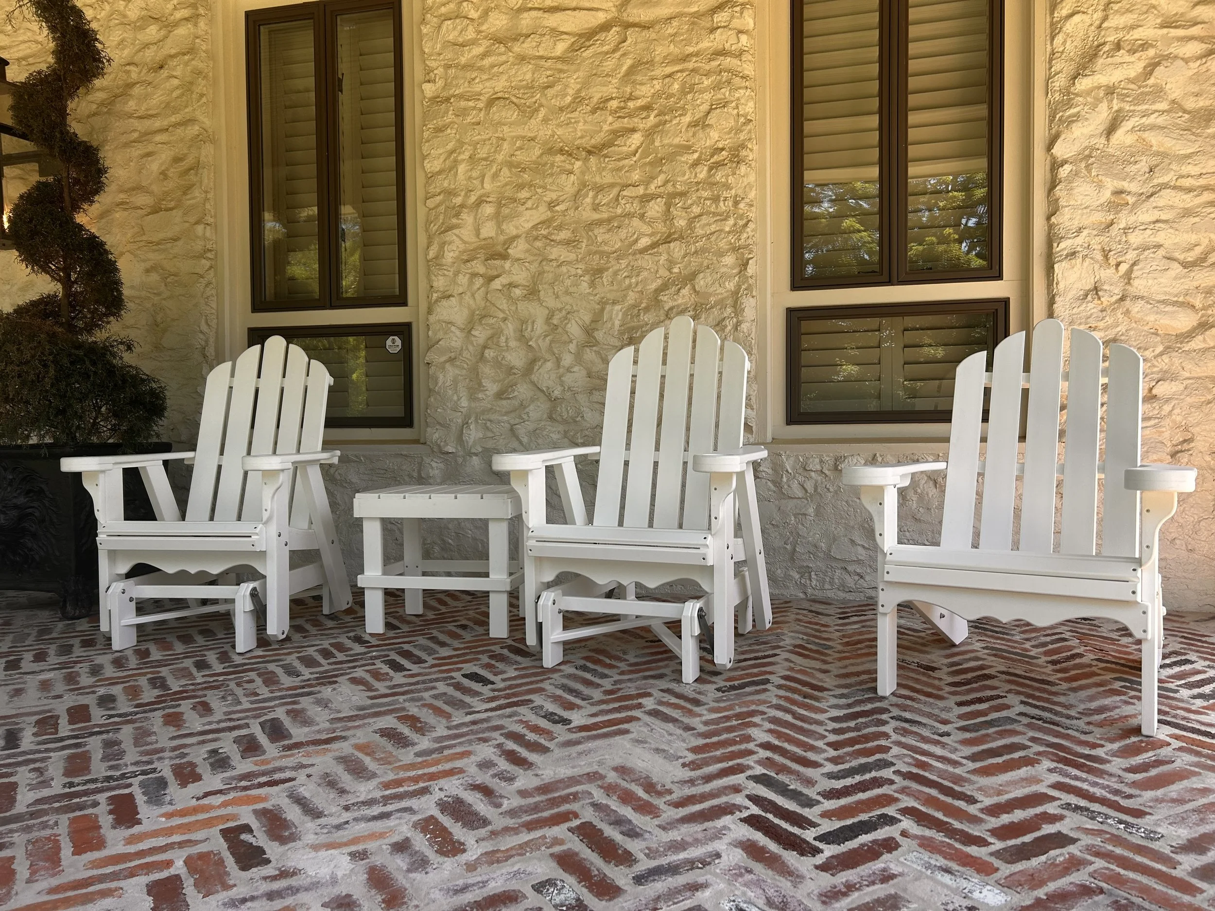 A row of white Adirondack chairs on a brick patio in front of a beige stone house with four black-framed windows.
