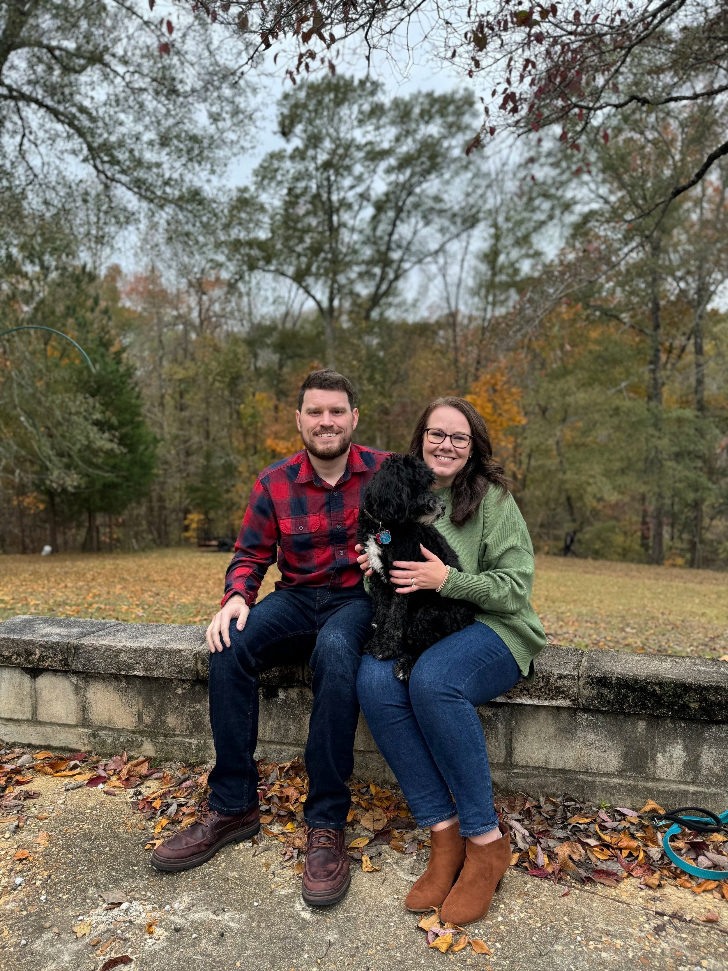 A smiling couple sitting on a stone bench outdoors in a park with autumn foliage, holding a black dog. The man is wearing a red plaid shirt and dark jeans, while the woman is wearing a green sweater and brown boots. Fallen leaves are scattered on the ground.
