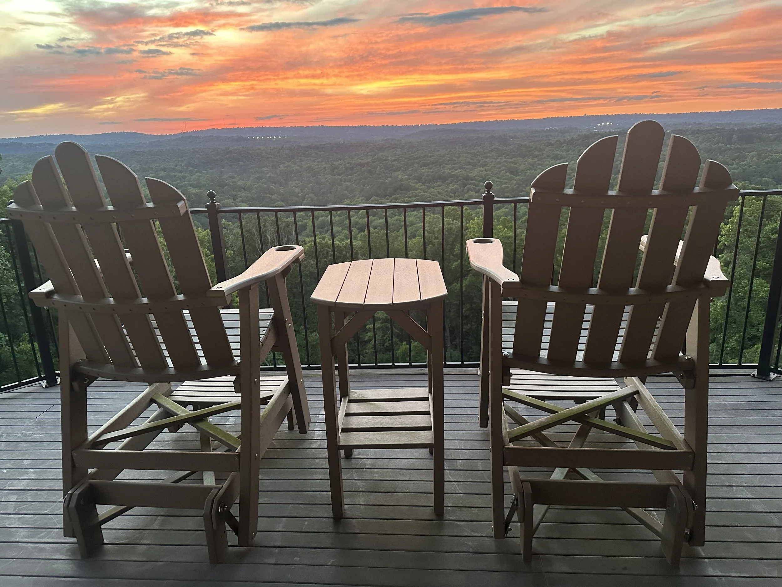 Two wooden Adirondack chairs and a small wooden table on a balcony with a view of a forest and a colorful sunset sky.