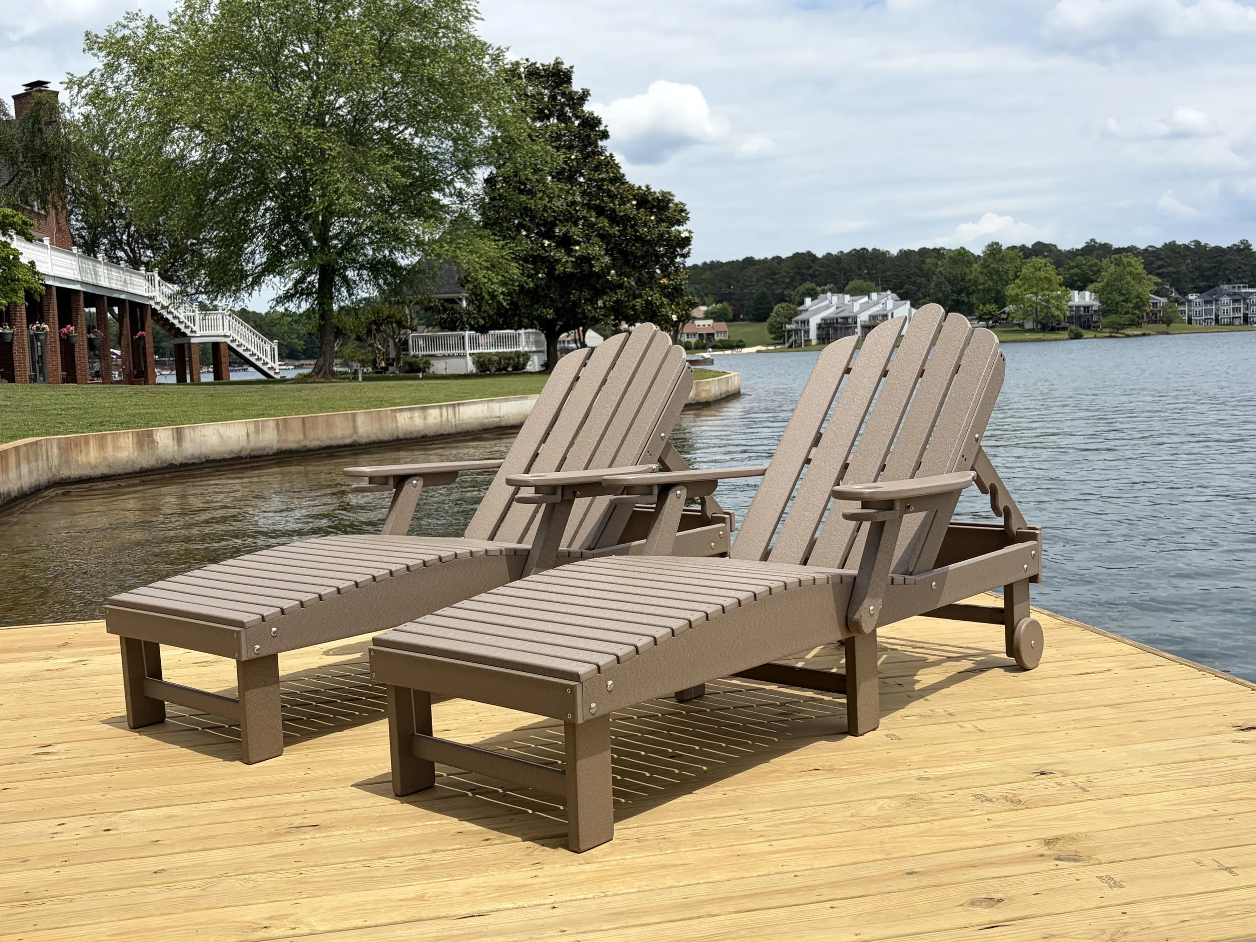 Three beige Adirondack chairs on a wooden deck by a lake with trees and houses in the background.