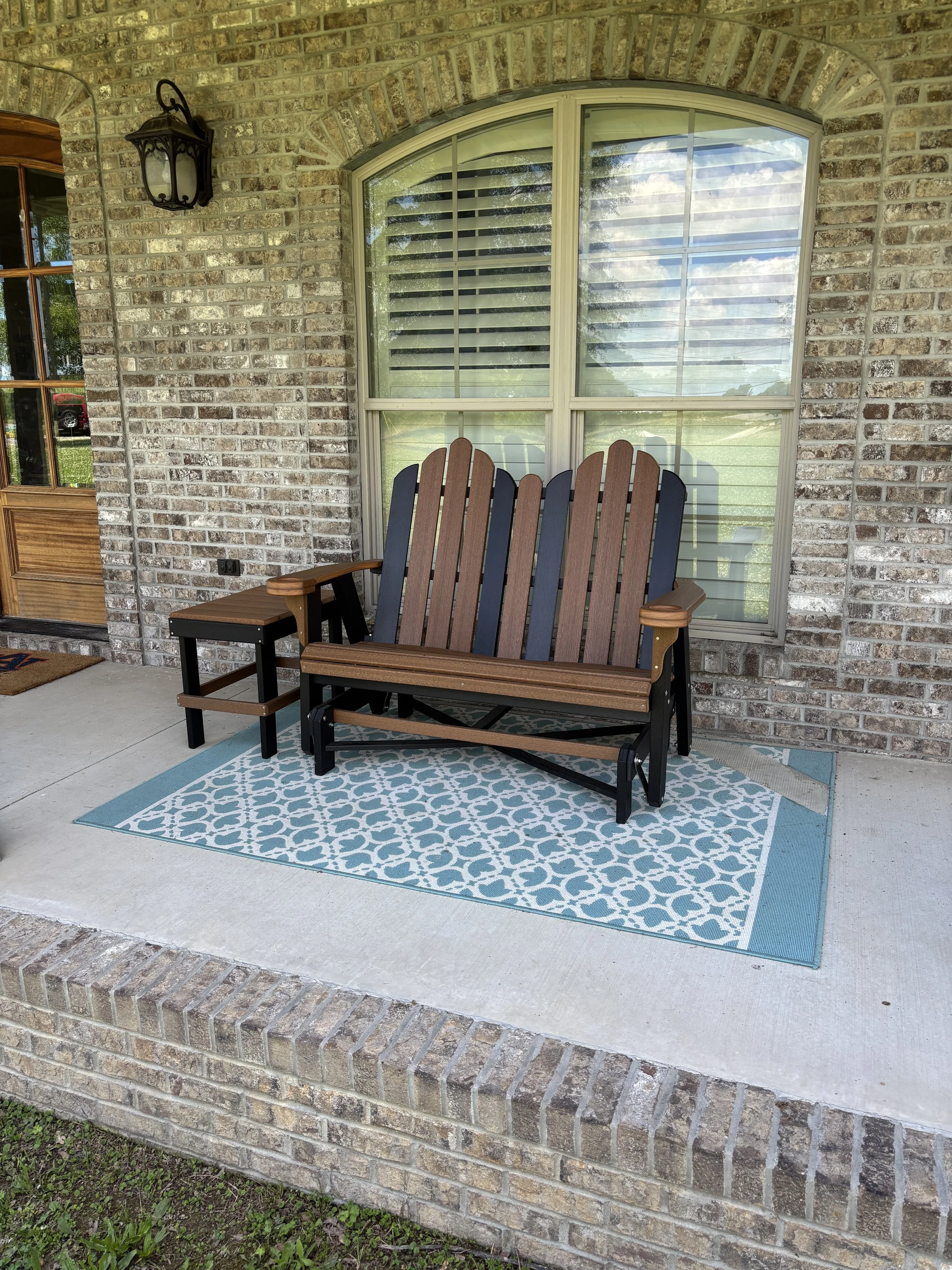A porch with a brick wall, a large window with shutters, a wall-mounted lantern, a wooden bench with metal armrests, and a blue and white patterned outdoor rug.