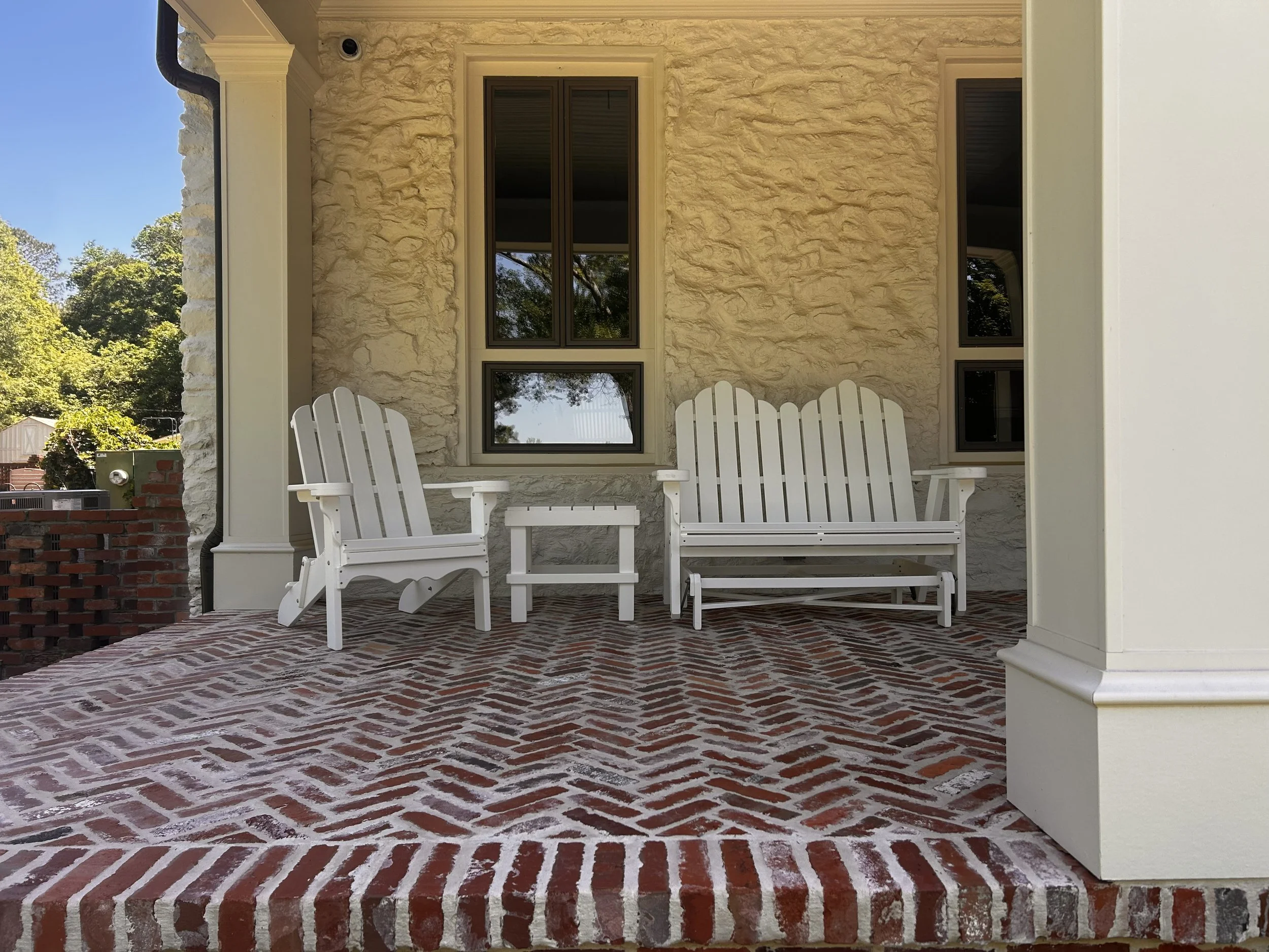 Front porch with brick flooring, white Adirondack chairs, textured beige stone house wall with three windows, and green trees in the background.
