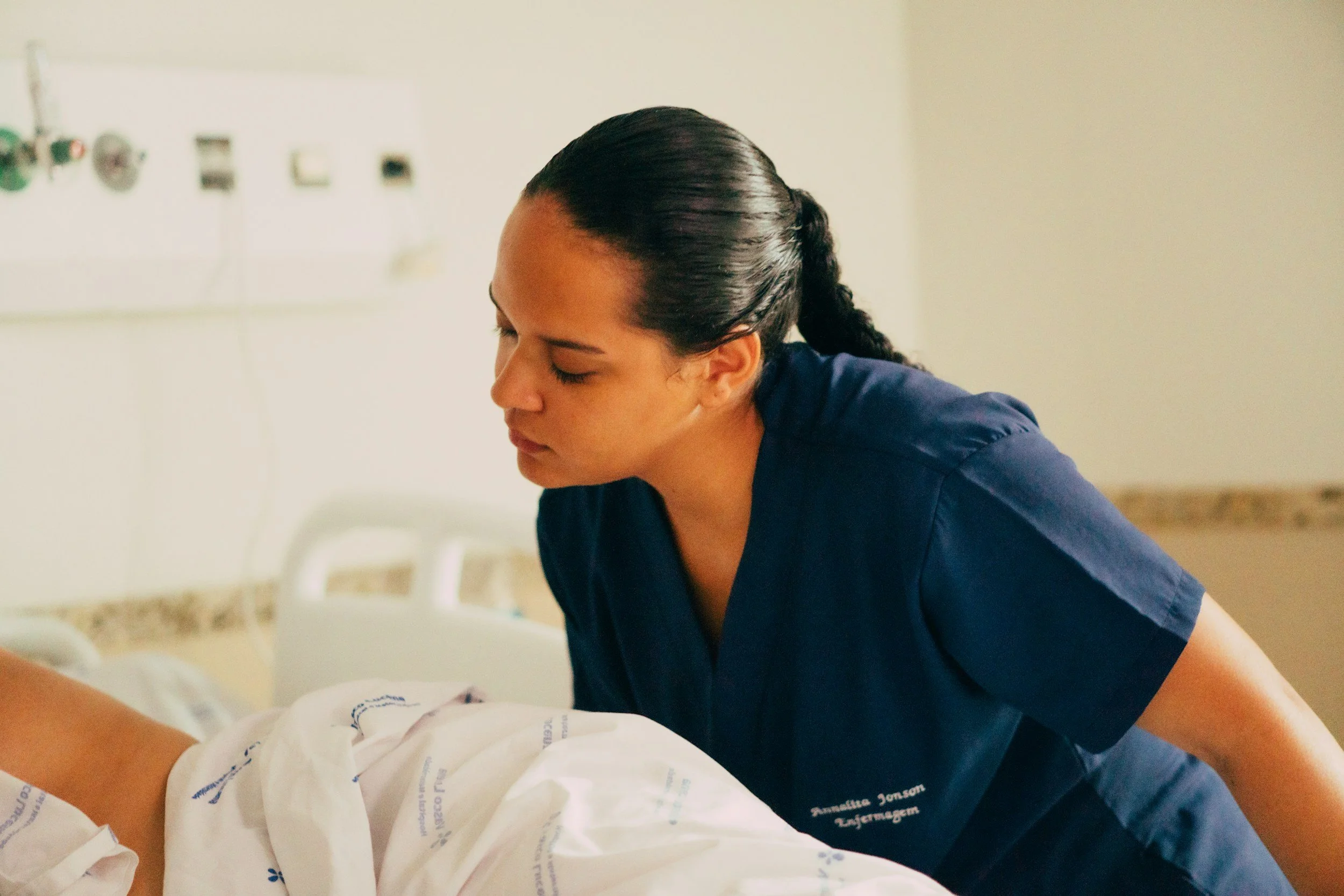 Dark-haired new grad NP in medical scrubs leaning over a patient in a hospital bed, working her dream job