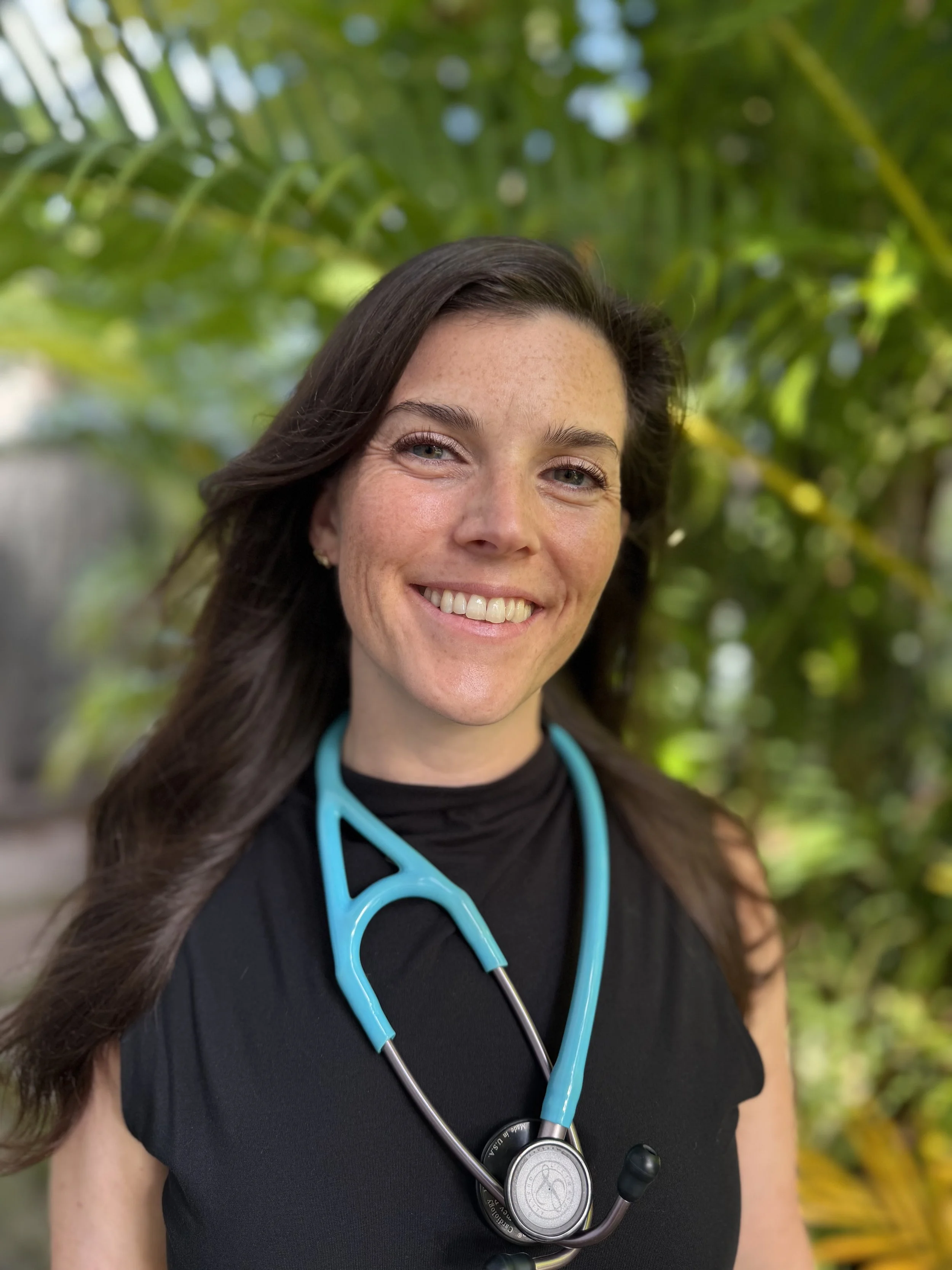 Close-up of a smiling woman wearing a black shirt with a blue stethoscope around her neck, outdoors with green foliage in the background.