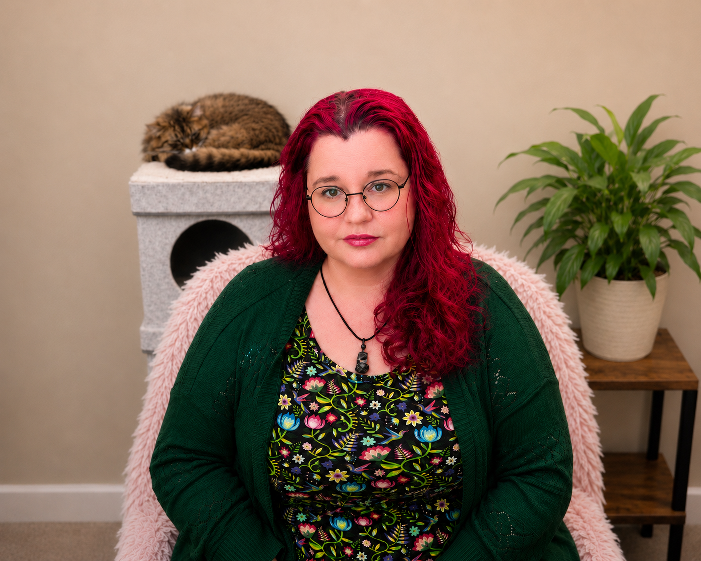 A woman with bright red curly hair and glasses sitting on a pink fluffy chair, with a tabby cat sleeping on a beige cat tree in the background to her left and a green potted plant on a small wooden table to her right.
