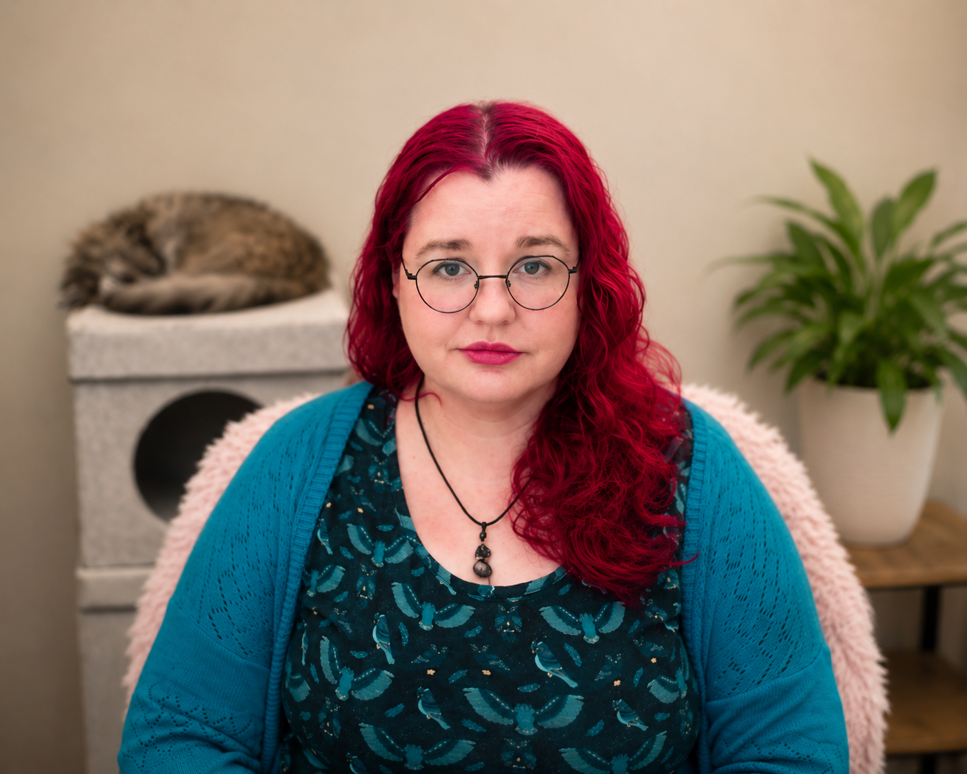 A woman with red curly hair, glasses, and a blue cardigan sitting in front of a beige wall, with a cat sleeping on a white cat condo and a potted plant in the background.