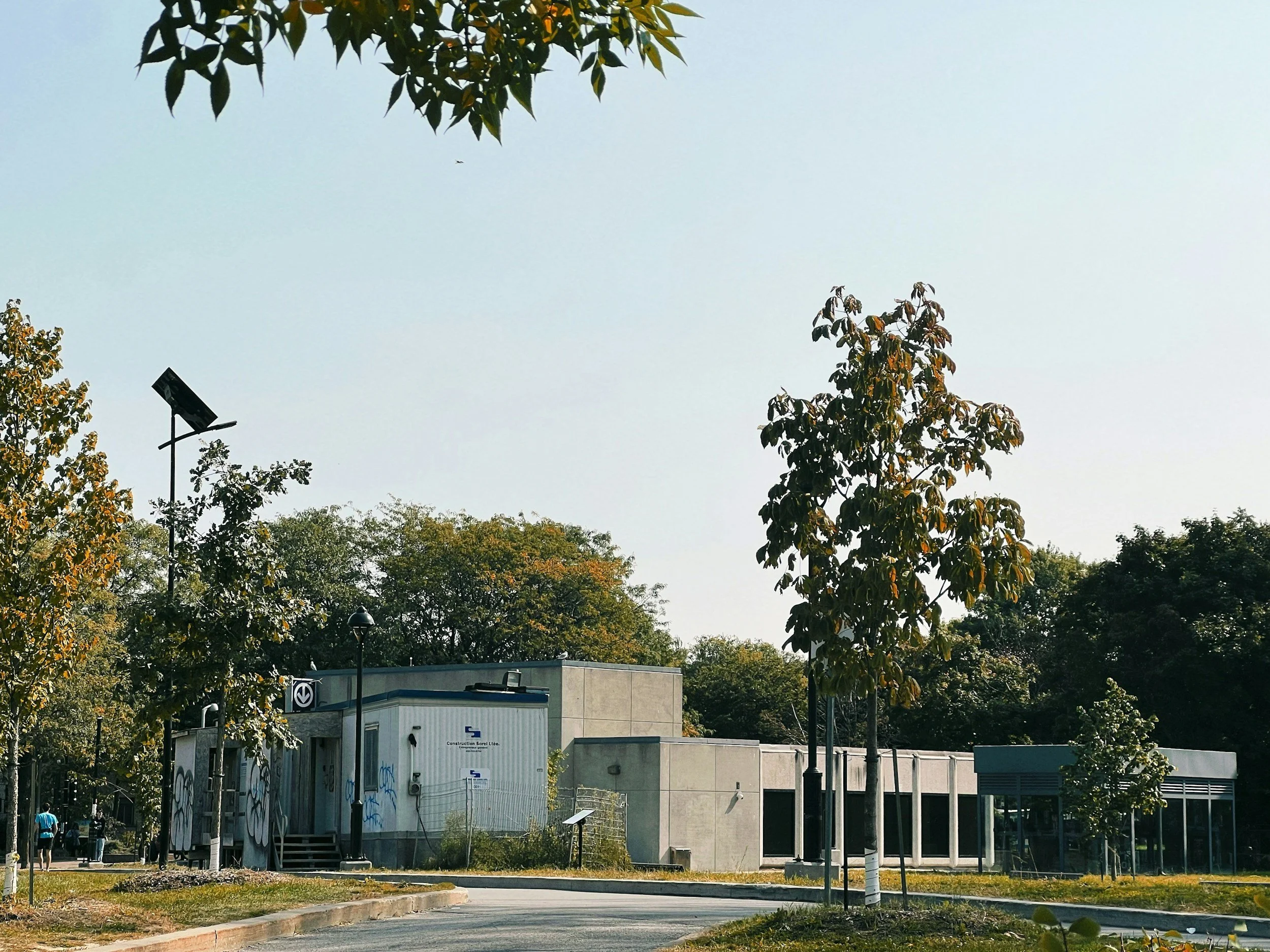 A street scene with trees, a modern building, streetlights, and people walking. There is a solar panel on a pole and some graffiti on the structure.