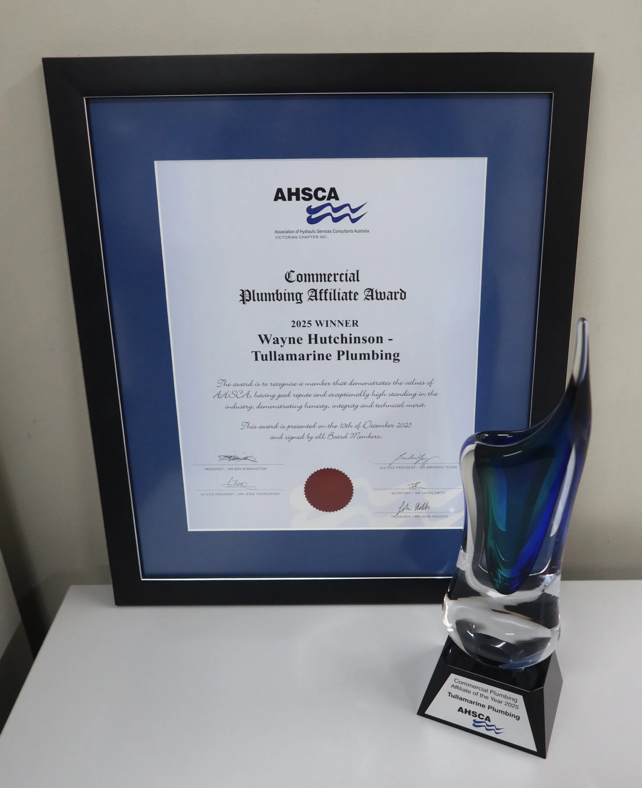 Certificate of award for Commercial Plumbing Affiliate of the Year 2025 presented to Wayne Hutchinson from Tullamarine Plumbing, awarded by AHSCA, displayed in a black frame on a wall, with a glass sculpture with blue, black, and clear swirls placed beside it on a white surface.