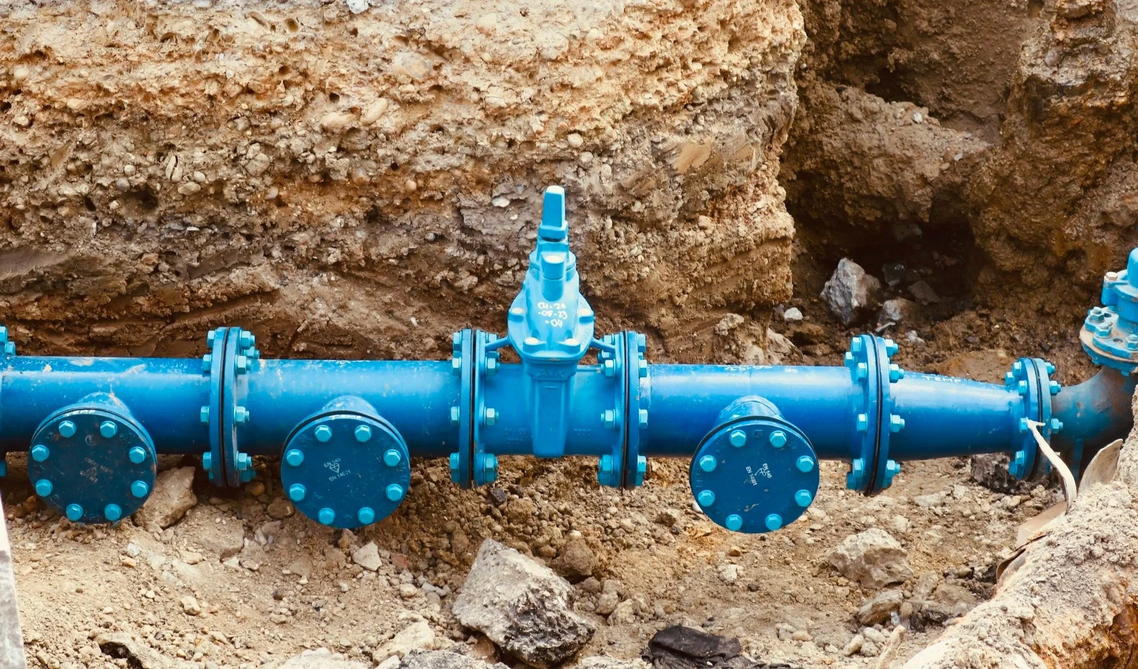 Blue water valve connected to a pipeline in a dug-up construction site with soil and rocks.