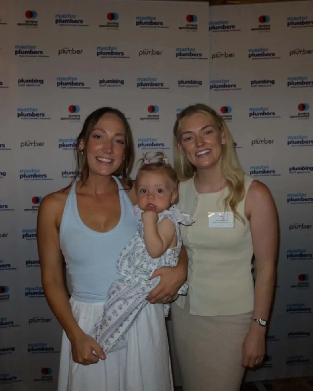 two women and a baby standing in front of a sign at a master plumbers event wearing a blue singlet top and white skirt and the baby is wearing a beautiful matching set of white and blue and flowers