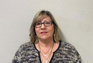 A middle-aged woman with glasses smiling against a wall wearing golf jewellery and an open cardigan against the wall in her office