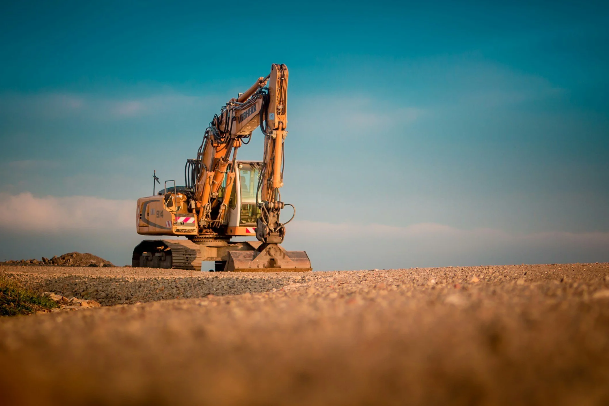 A large yellow excavator working on a construction site, with a clear blue sky in the background and gravel on the ground.