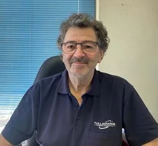 A middle-aged man with short gray hair and gray beard sitting on a beige bench against a plain, light-colored wall. He is wearing a brown polo shirt and light-colored trousers, looking thoughtfully to the side.