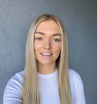 A young woman with blonde hair, wearing a blue sweater and beige pants, sitting on the edge of a beige bench with his hand resting on the surface, looking to the side. The background has a plain wall with soft shadows.