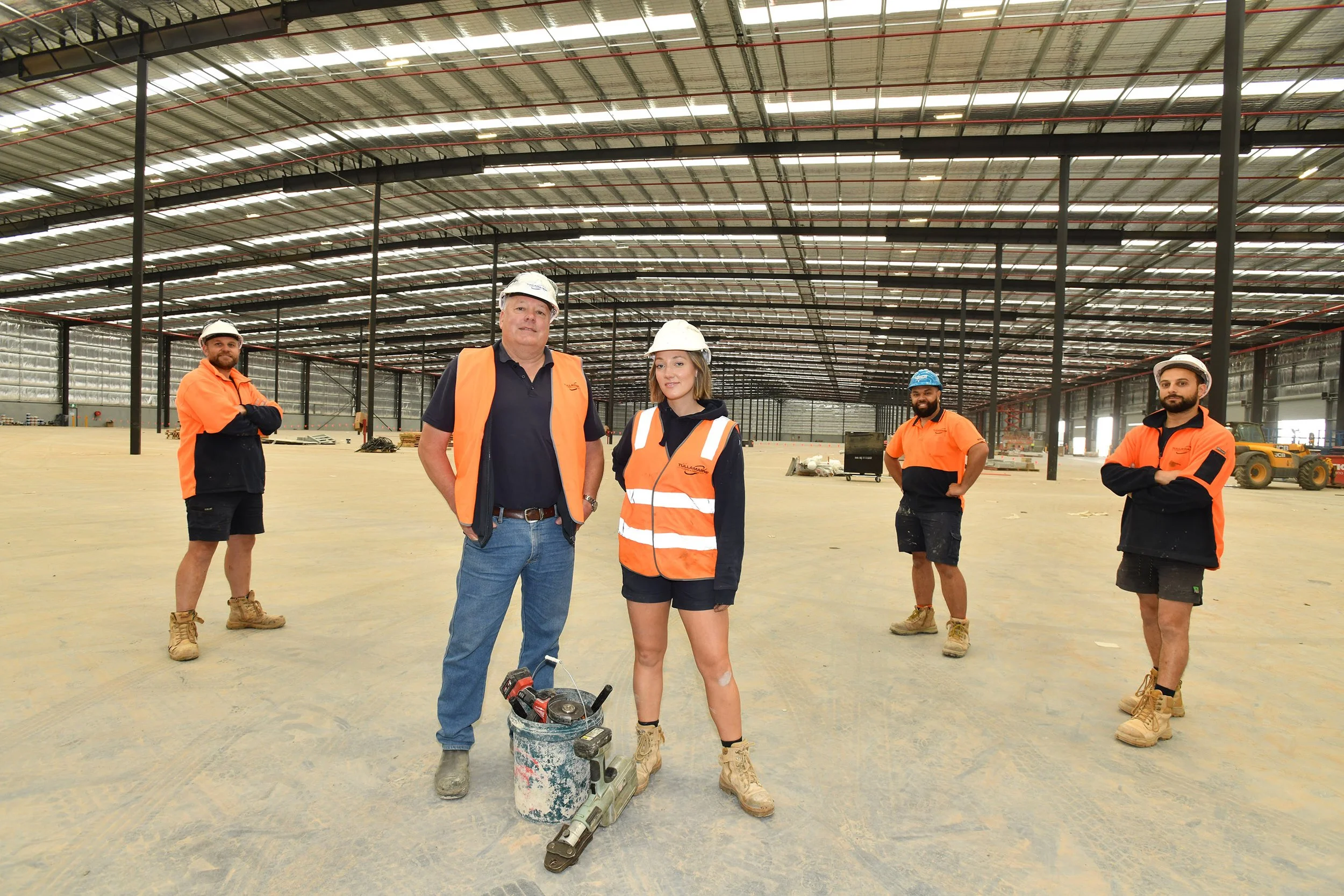 Group of five construction workers in an unfinished large indoor building, wearing safety helmets and orange safety vests, with one person in a black polo shirt, holding a power tool, and a bucket with construction tools on the floor.