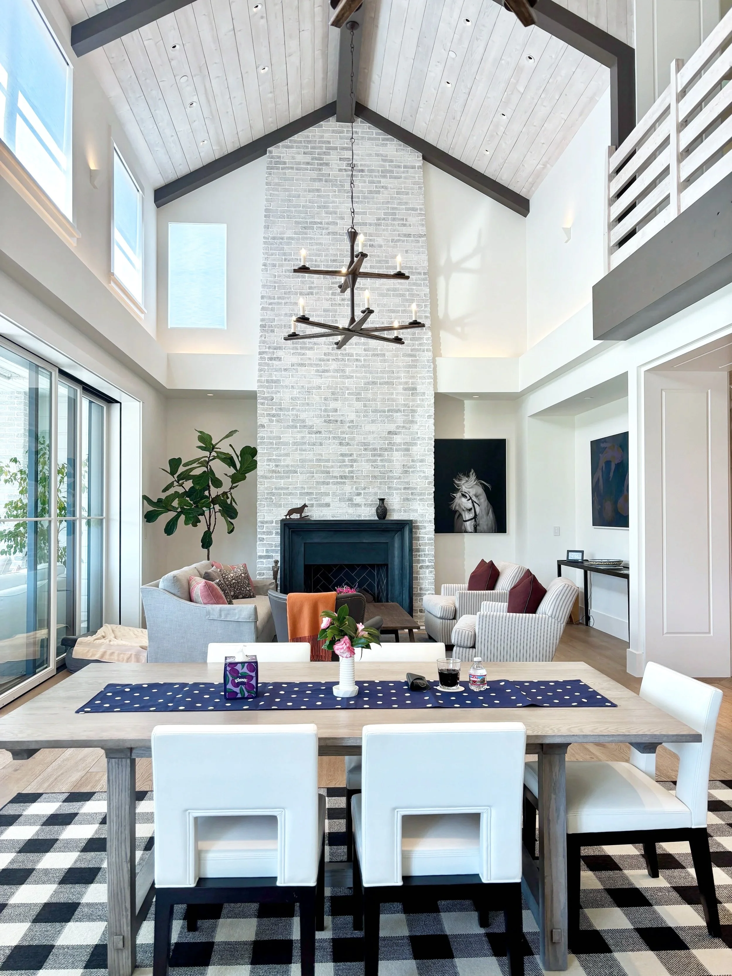 Interior of a modern living room with high vaulted ceiling, white brick chimney, sofas, artwork, and dining area, near large windows.