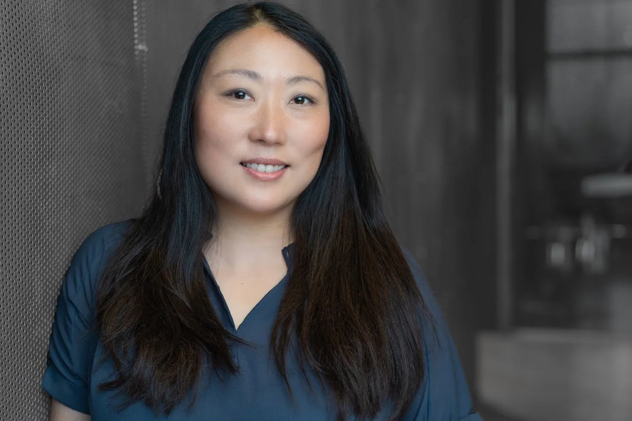 Portrait of a woman with long dark hair smiling, wearing a blue top, standing beside a textured wall in a professional setting.