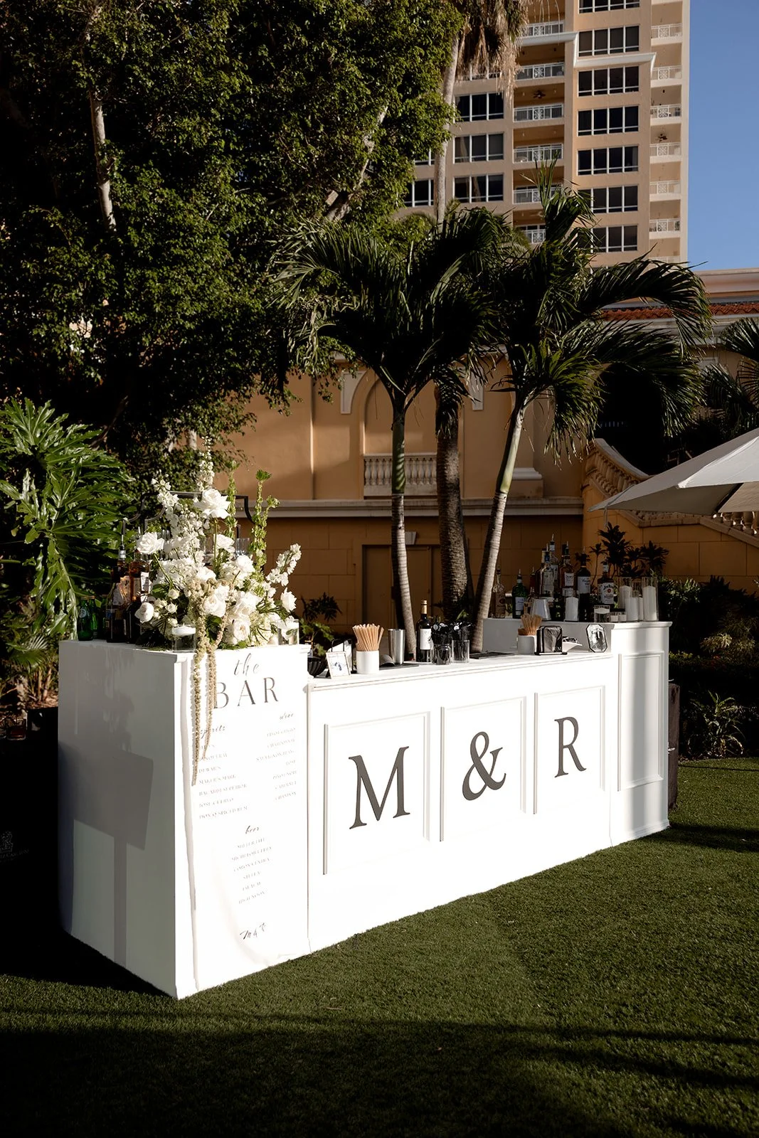 Outdoor wedding bar station with white paneling, decorated with white flowers and greenery, set against a backdrop of trees and a tall apartment building.
