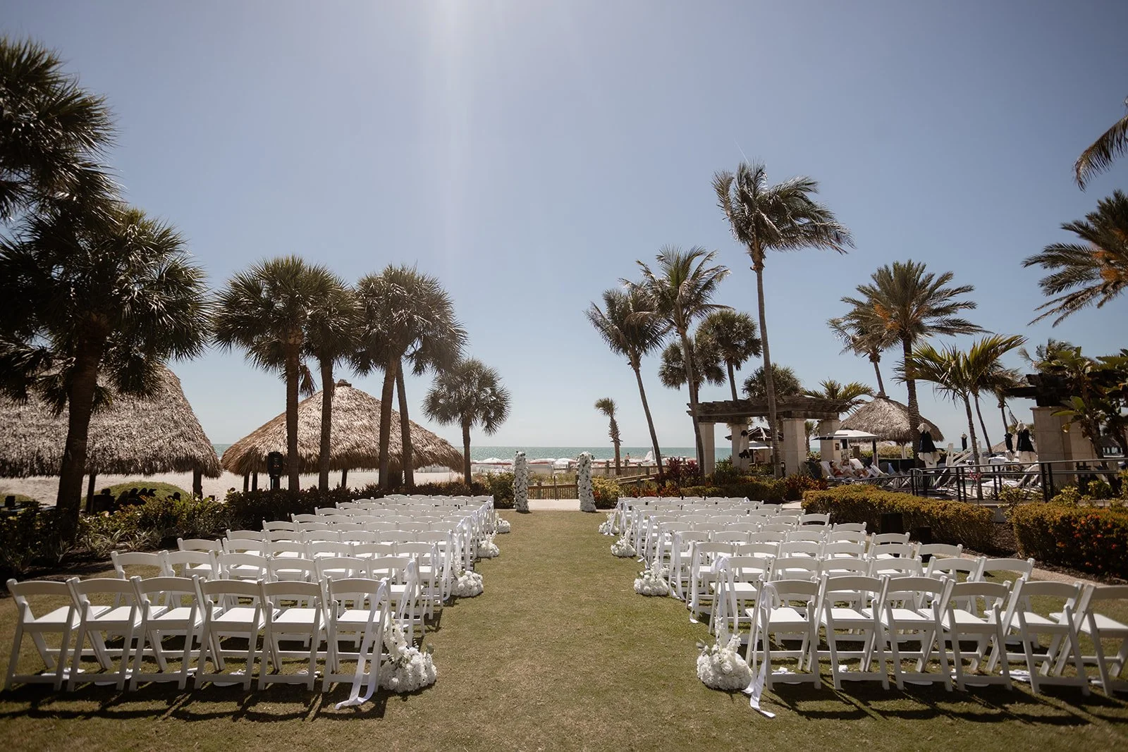 Outdoor wedding ceremony setup on a grassy area with white chairs, palm trees, thatched umbrellas, and ocean in the background on a sunny day.