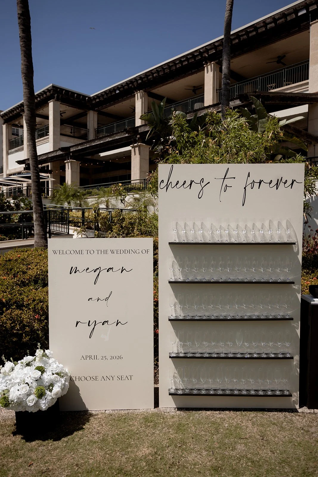 Wedding welcome sign with the names Megan and Ryan and the date April 25, 2026, inviting guests to choose a seat, and a separate sign with the phrase 'cheers to forever' and empty glass shelves for drinks, outdoors with a building and palm trees in the background.