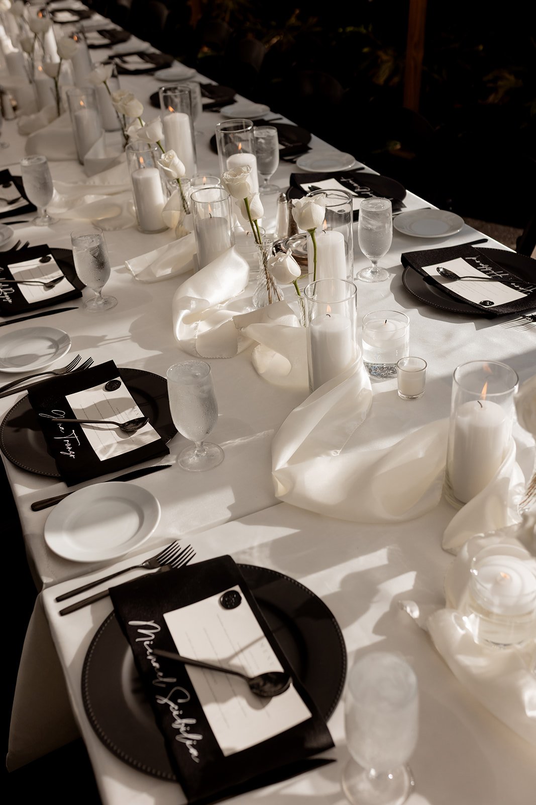 A long dining table decorated with white flowers, candles, black plates, and place cards, set for a formal event or dinner party.