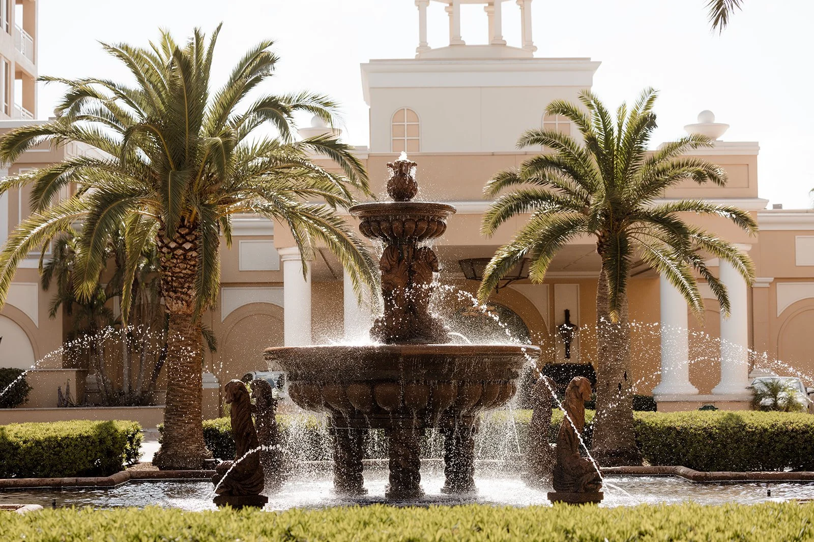 An ornate fountain with water streaming from multiple tiers, surrounded by palm trees and green bushes, in front of a beige building with white columns and a tower.
