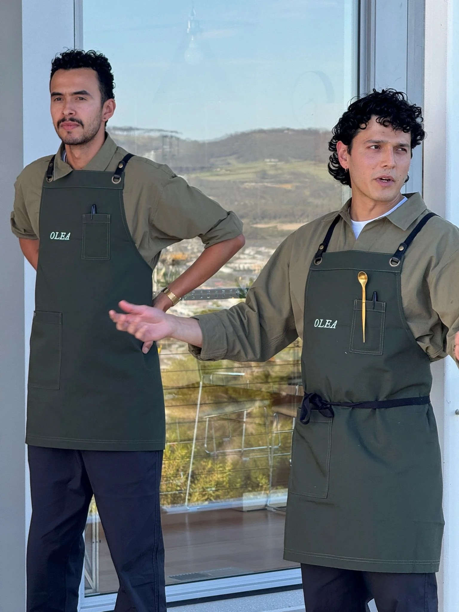Two male employees wearing khaki shirts and dark aprons with the name 'OLEA' printed on them, standing indoors near a large window with a scenic view of hills and sky in the background.