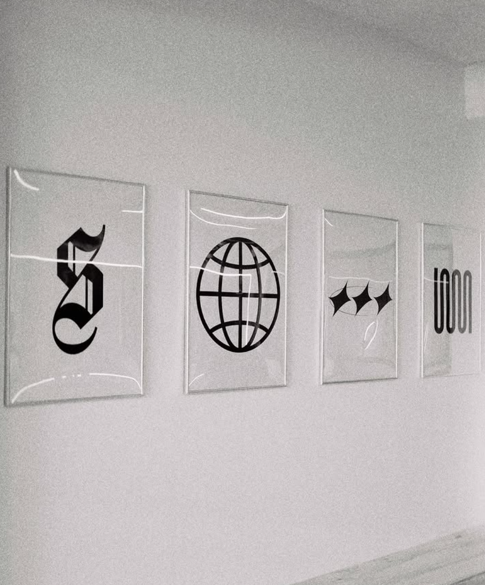 Four framed prints on a white wall with black symbols, including a stylized dollar sign, a globe, three diamond shapes, and a sequence of four vertical lines with the number 1000.