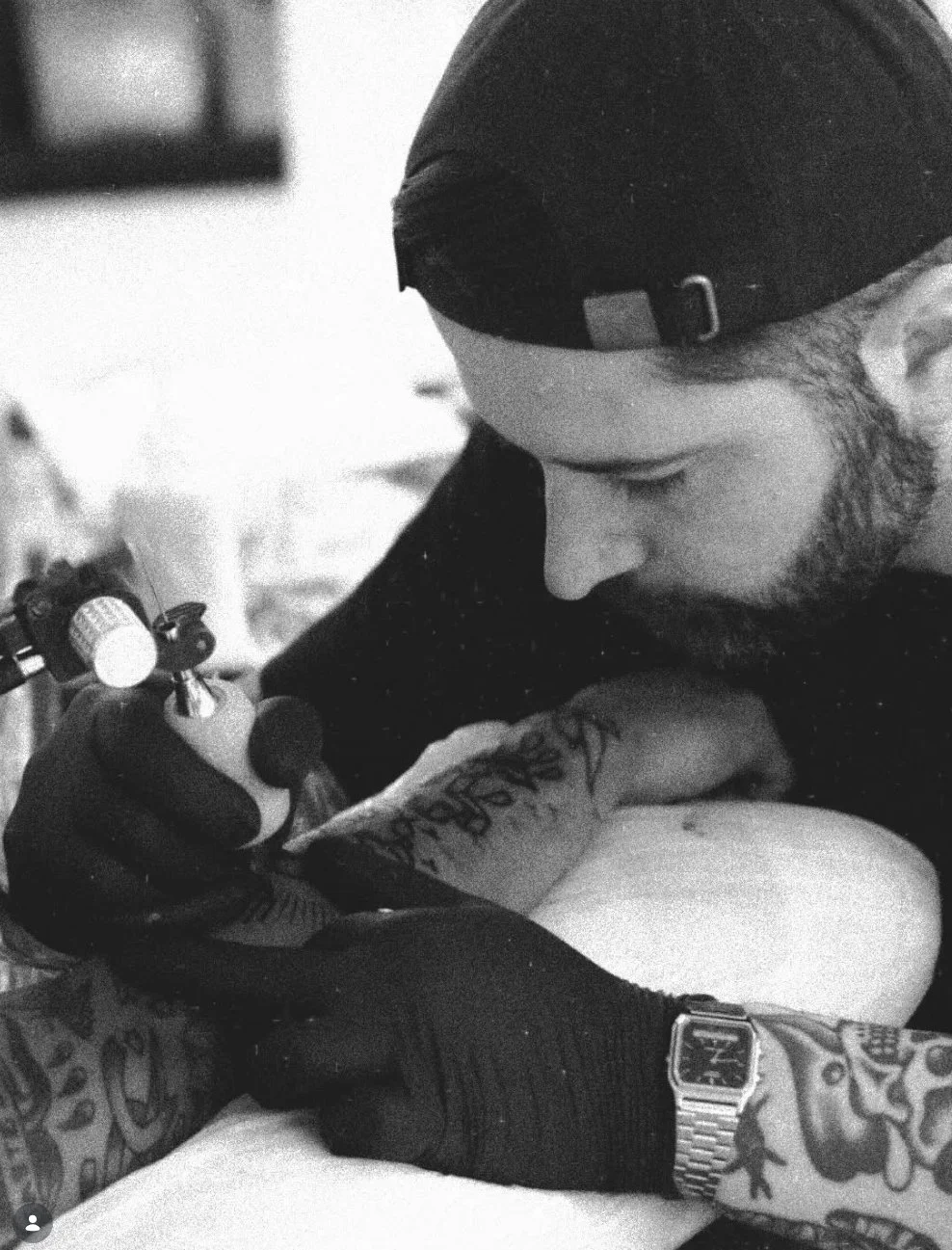 A person with a cap tattooed on their arm is tattooing another person's arm in a close-up, black-and-white photo.